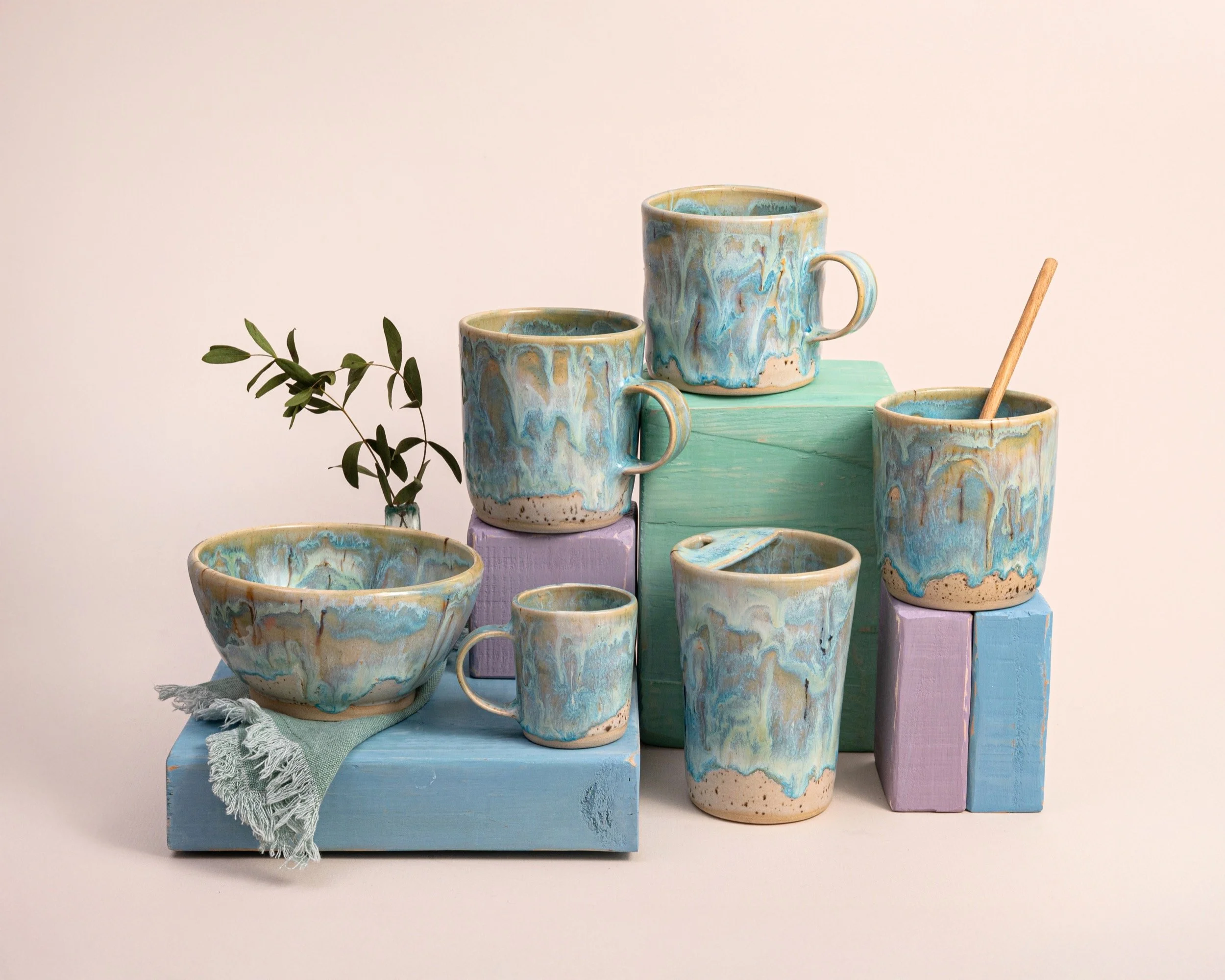 Ceramic mugs and bowls with blue, beige, and white marbled glaze on pastel-colored wooden blocks, with a small sprig of greenery.