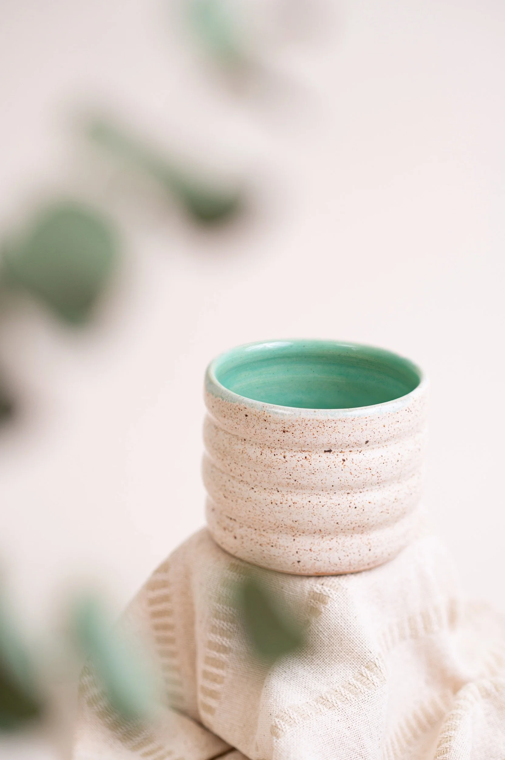 A textured ceramic cup with a turquoise interior sitting on a white cloth, with blurred green foliage in the background.