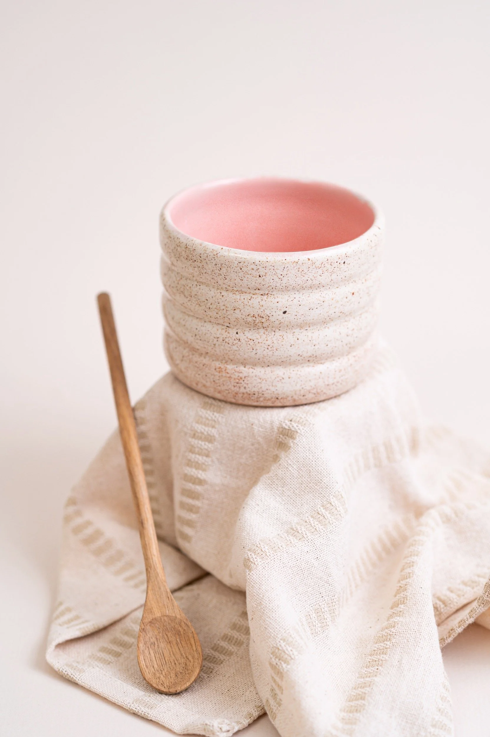 A pink and white speckled ceramic bowl with a pink interior, placed on a folded cream-colored cloth, next to a small wooden spoon on a neutral background.
