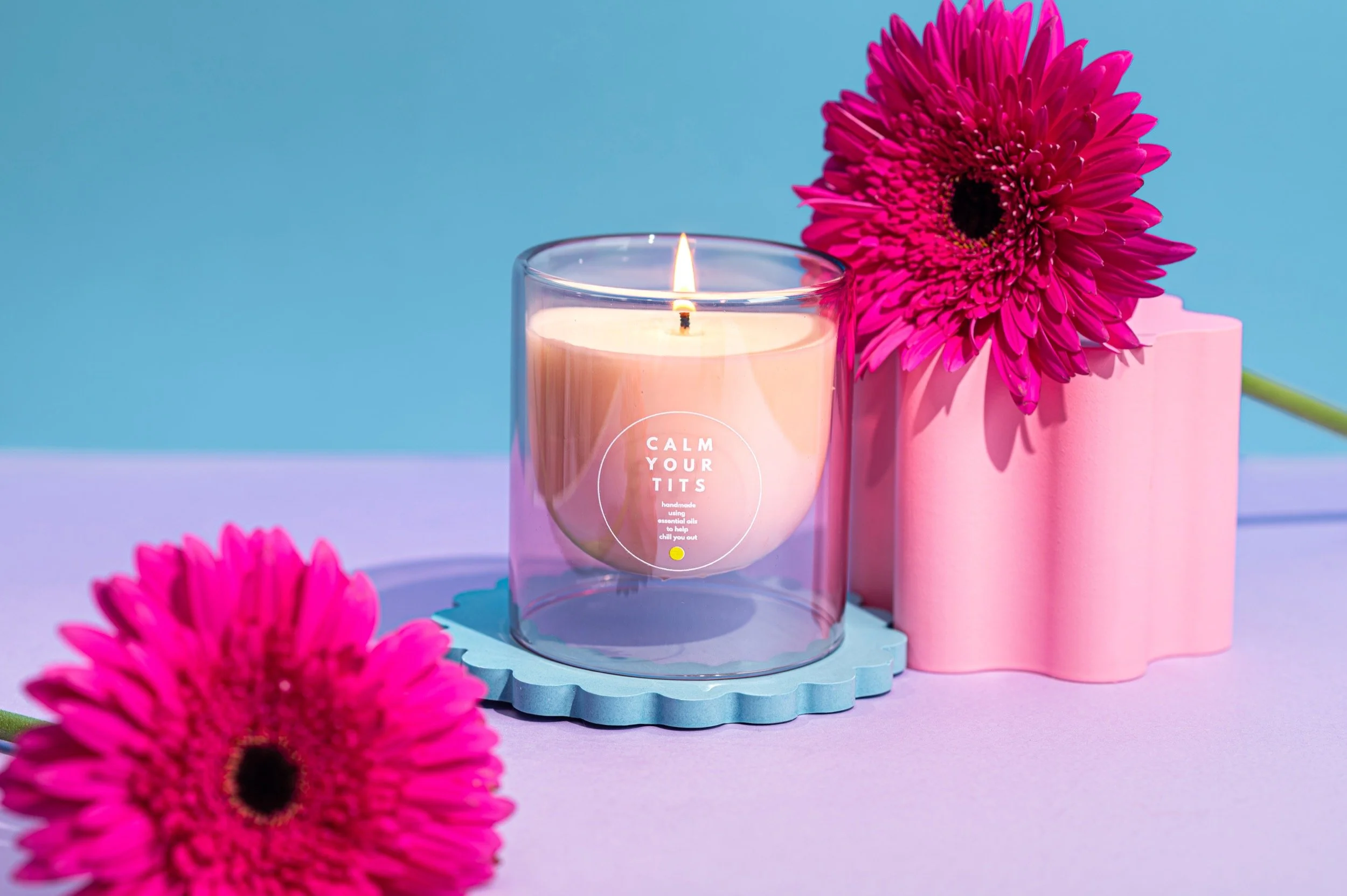 A glass candle sits on a scalloped disc with bright background and pink gerberas