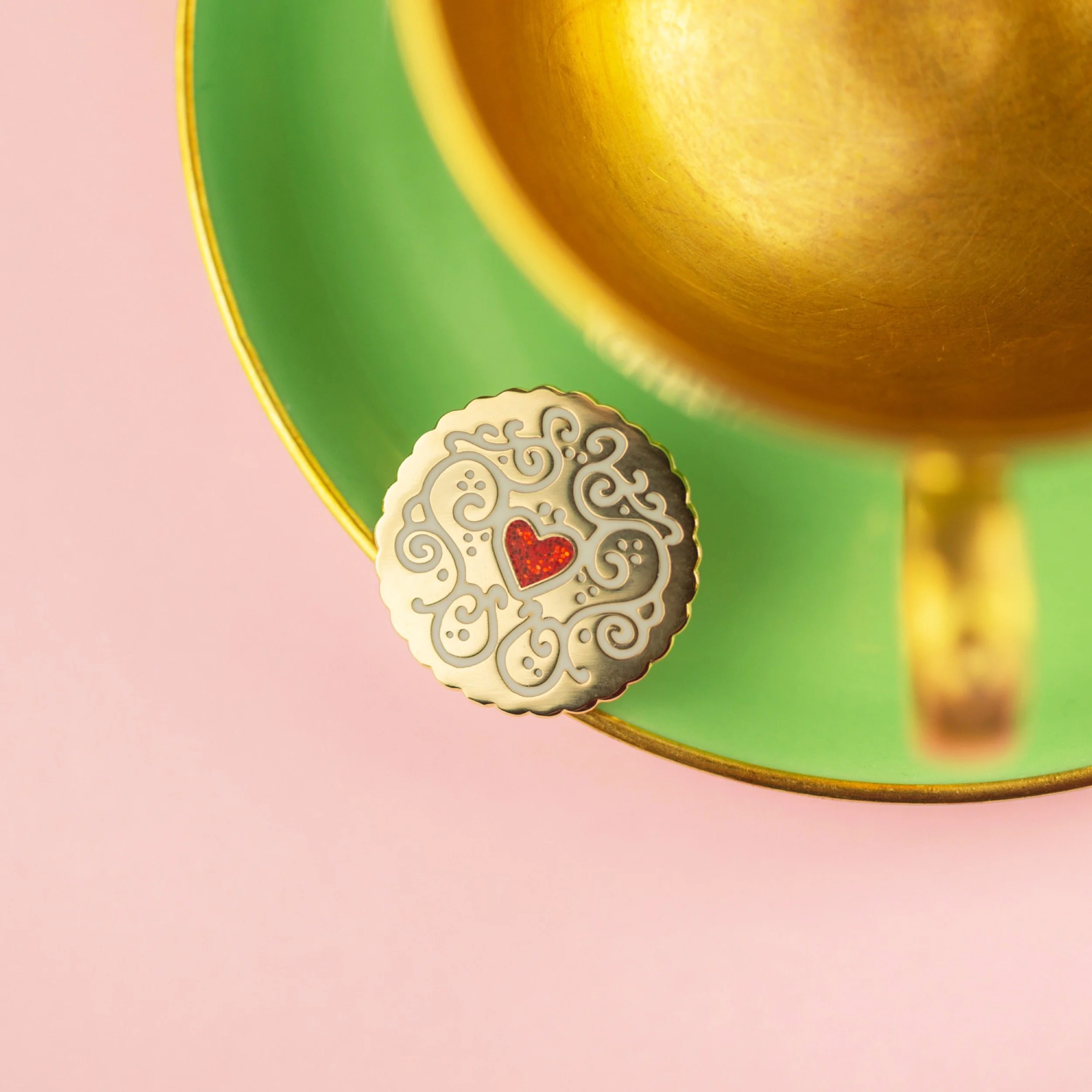 A jammy dodger enamel pin, resting on the edge of a green and gold vintage coffee cup against a pink background.