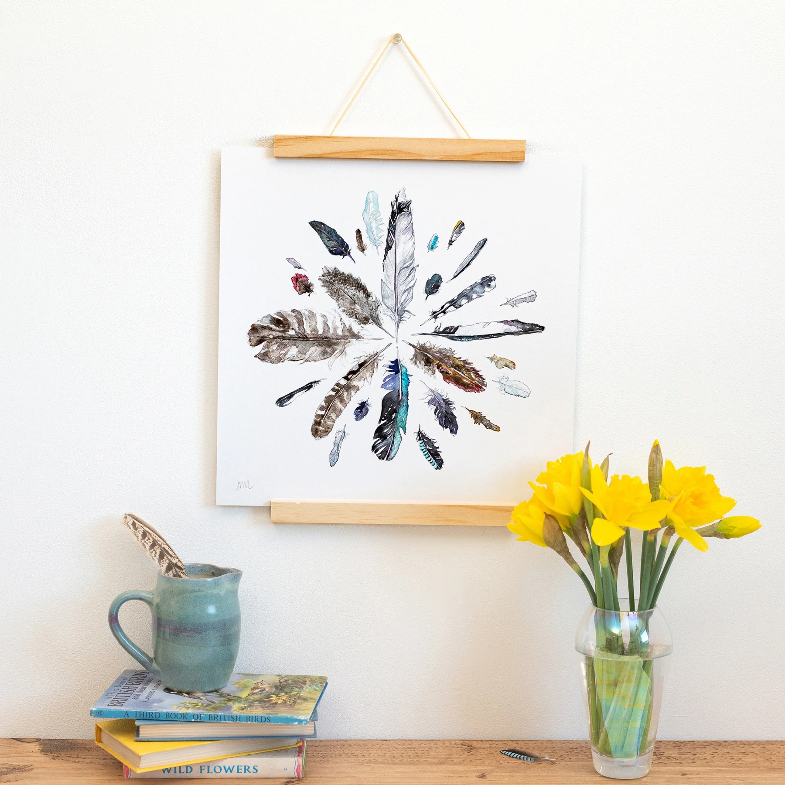 a feather illustration print hangs above a wooden shelf with books and daffodils