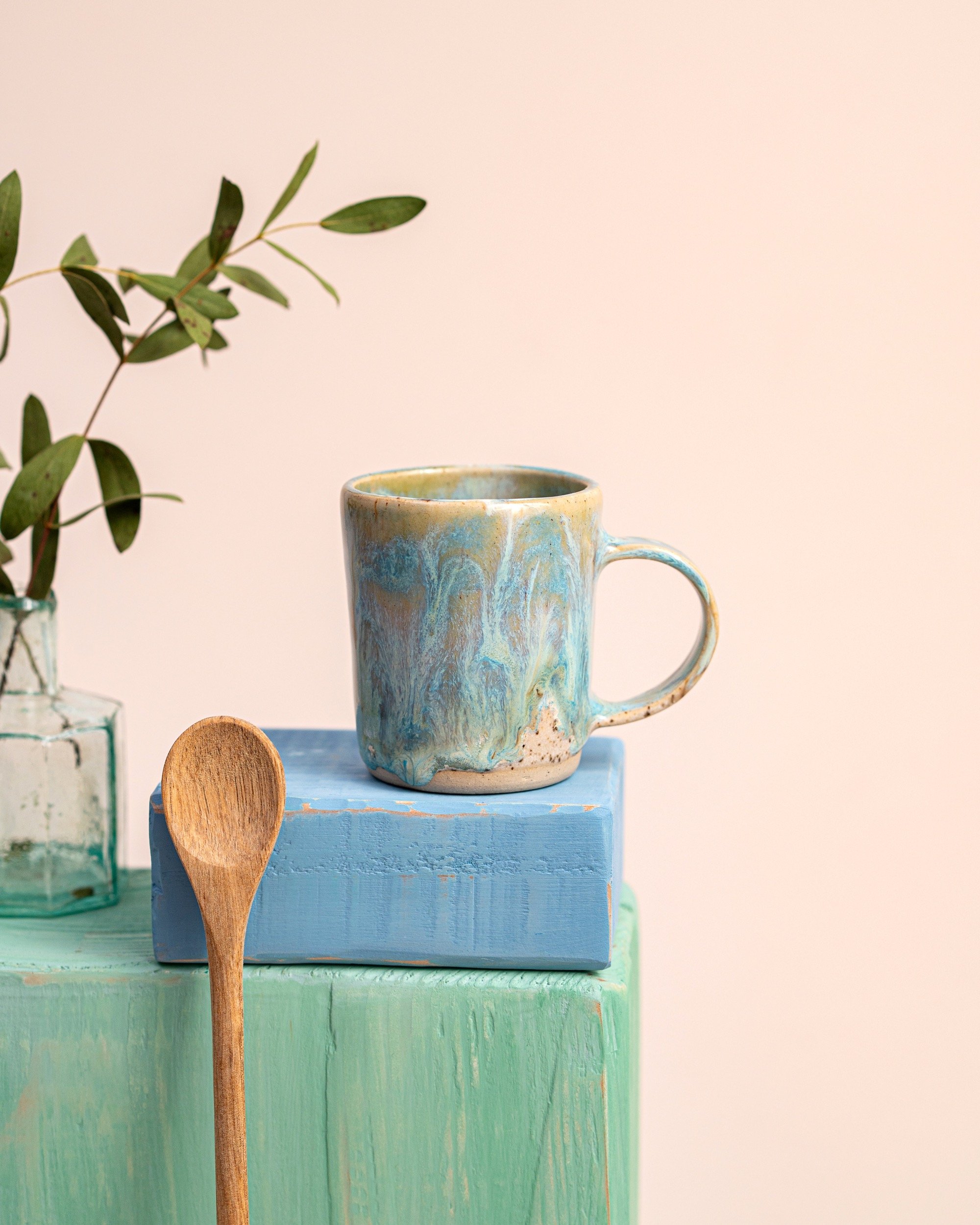 A marbled ceramic mug with blue, beige, and green swirls, placed on top of a painted blue wooden block. Next to it is a wooden spoon leaning against the block. To the left, there is a glass bottle with green leaves inside, against a plain light-color