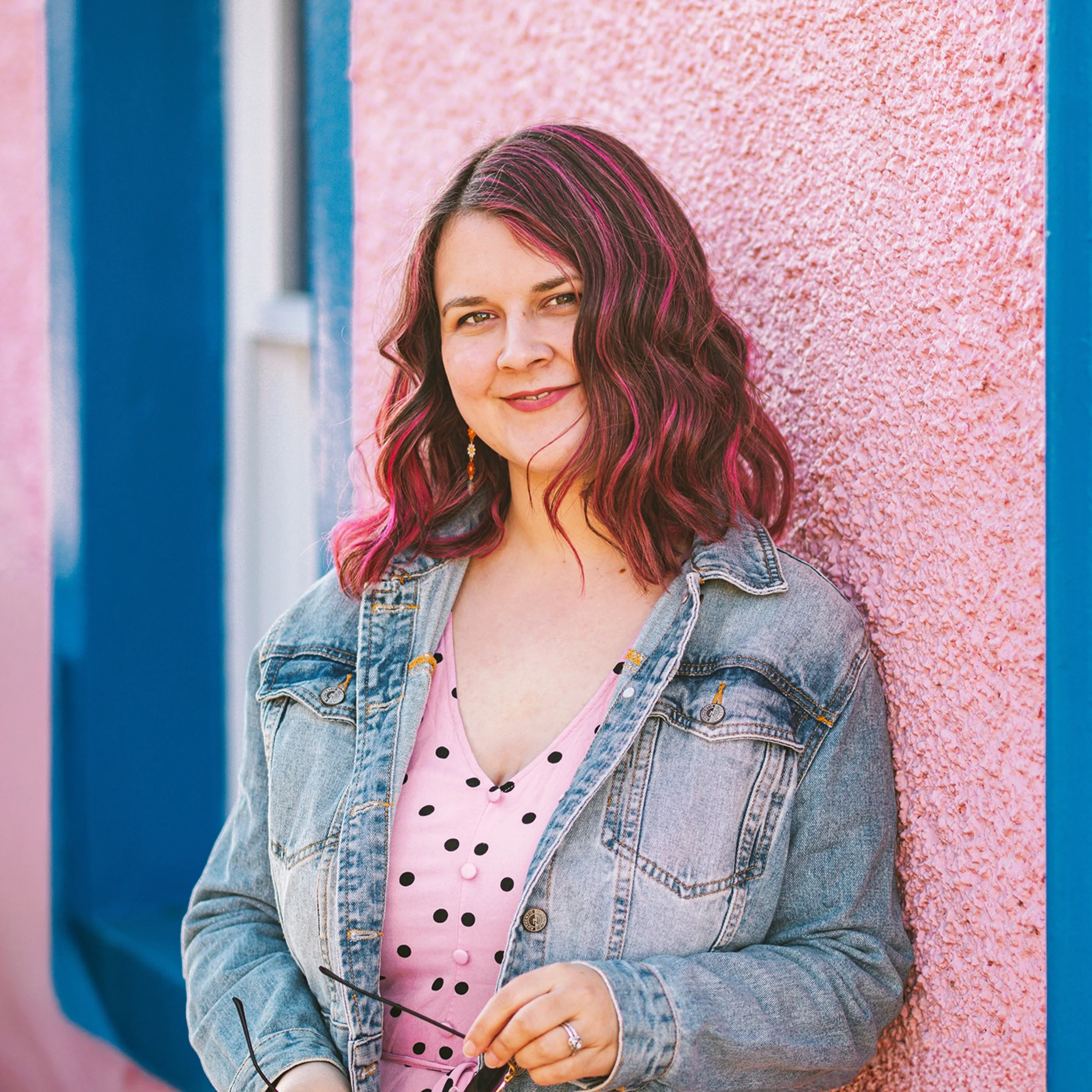 Jilly leans against a pink wall, she wears a denim jacket and has a pink dress on and pink in her hair