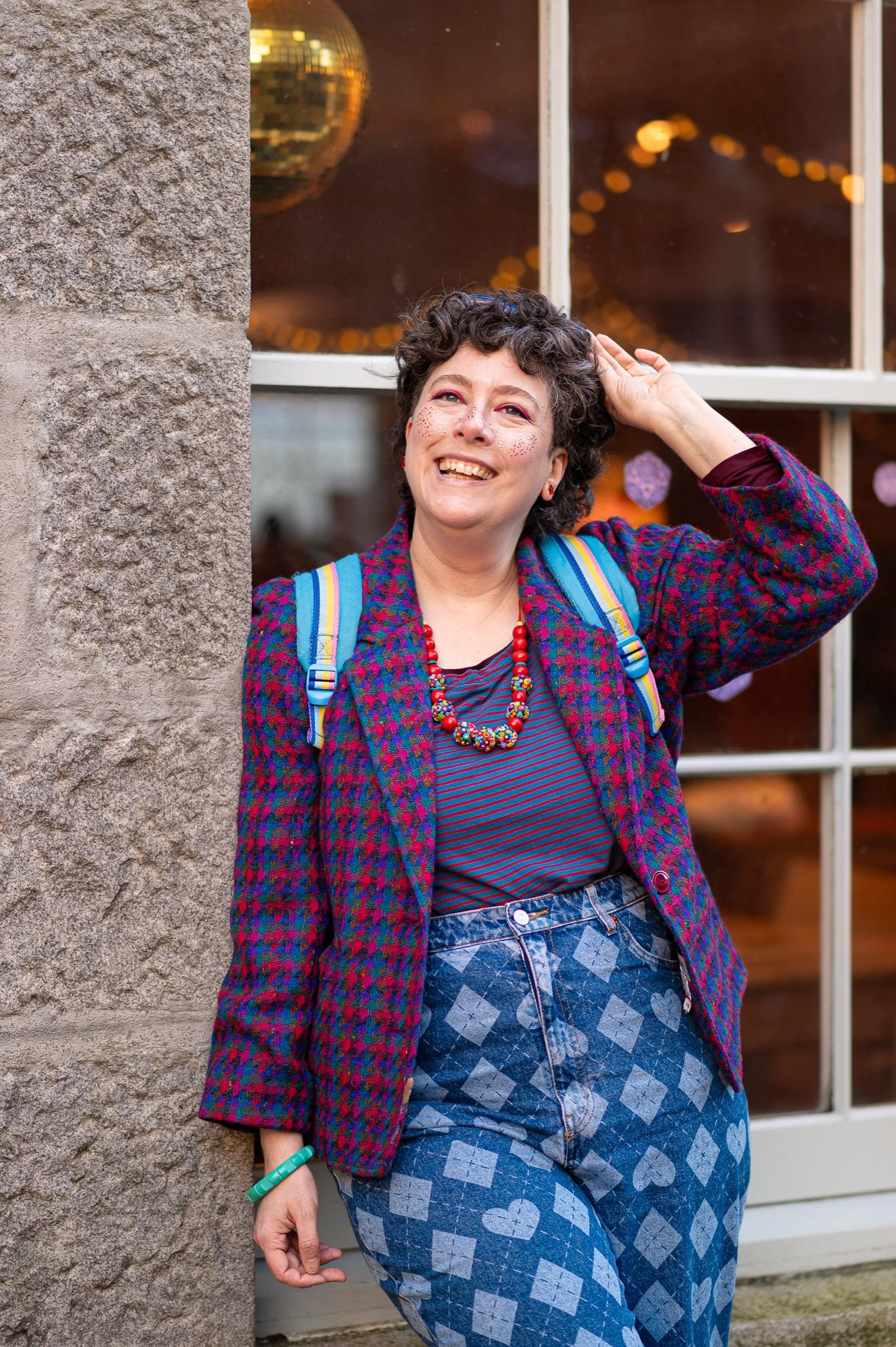 A smiling person with short curly hair, wearing colourful clothing and accessories, standing outdoors near a stone wall and window with reflections of bokeh lights.
