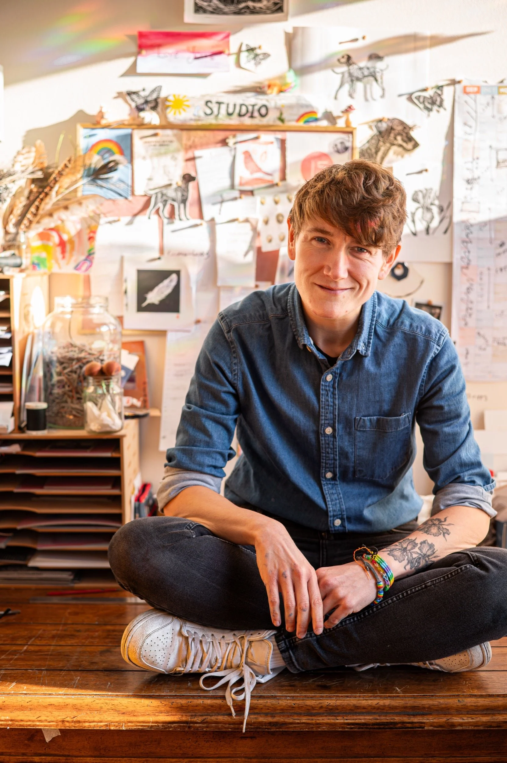 A woman sitting cross-legged on a wooden desk in a creatively decorated room, smiling at the camera. The background features a wall covered with drawings, notes, and crafts, along with a glass jar filled with art supplies.