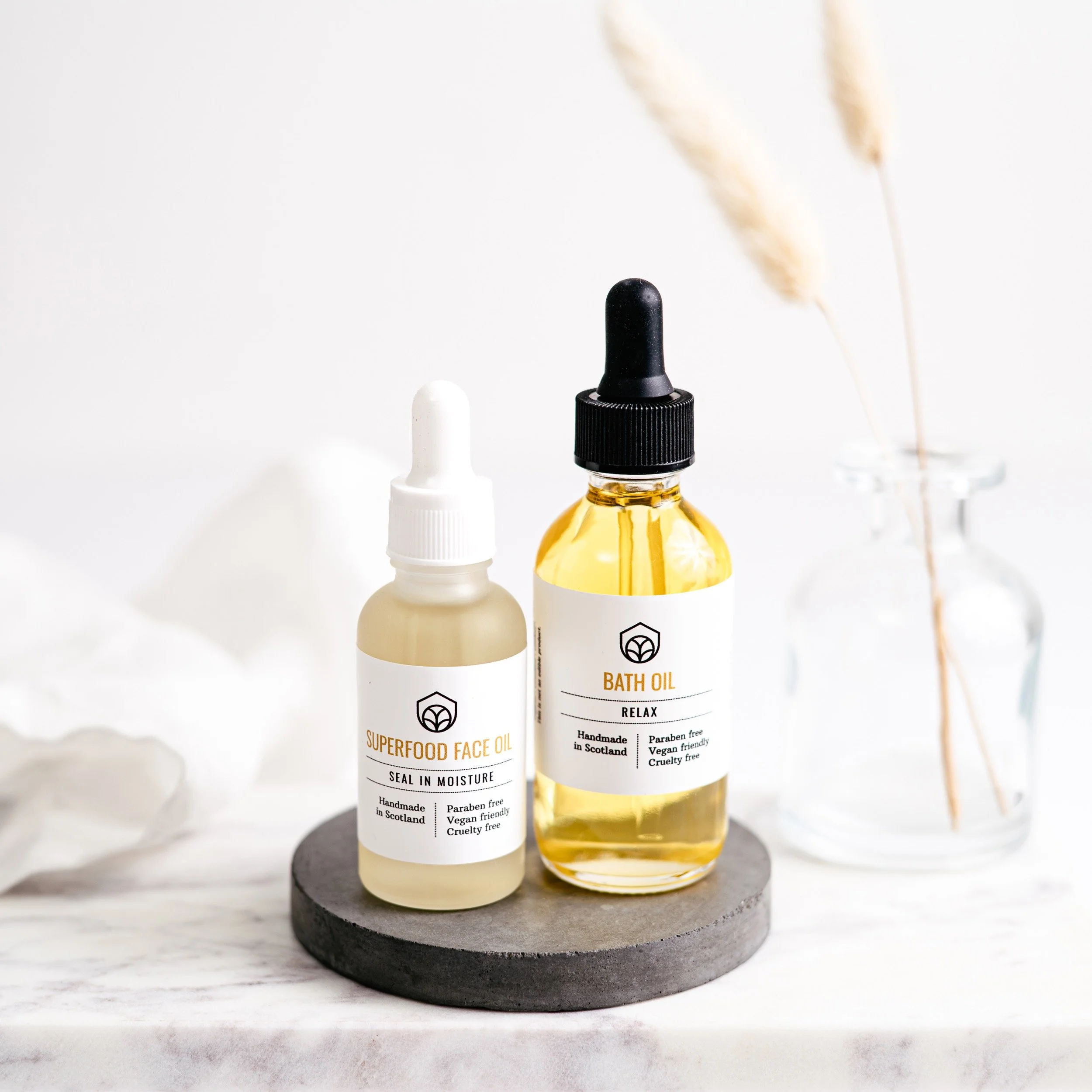 Bottles of skincare oils, one labeled Superfood Face Oil and the other Bath Oil, placed on a round black stand with a white marble surface, with a glass vase containing dried grass in the background.