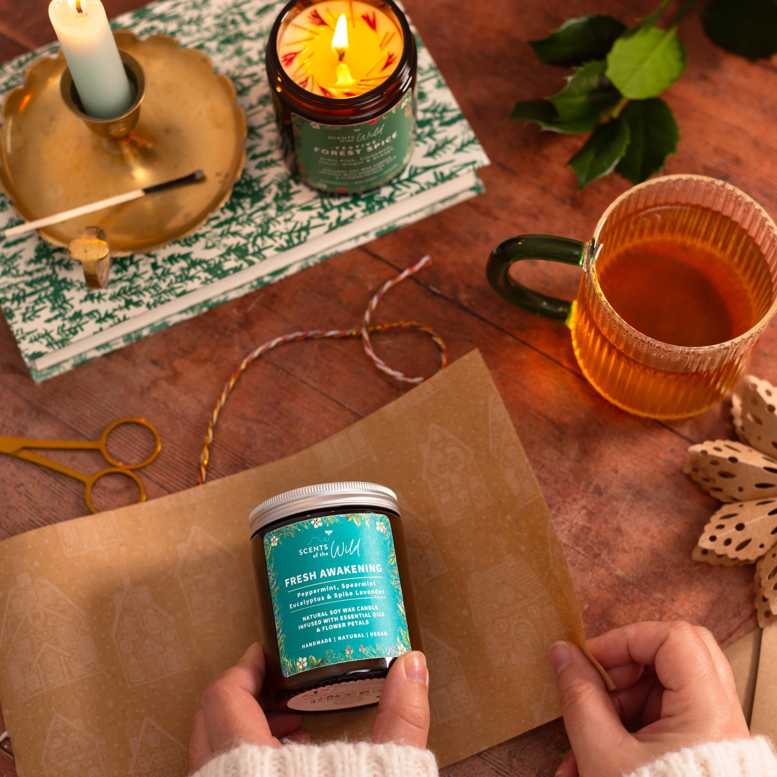 A person holding a jar candle labeled 'Fresh Awakening' with peppermint, spearmint, eucalyptus, and spike lavender scents. The table has a candle, a cup of tea, wrapped gifts, scissors, a candle holder with a lit candle, and a decorative butterfly or