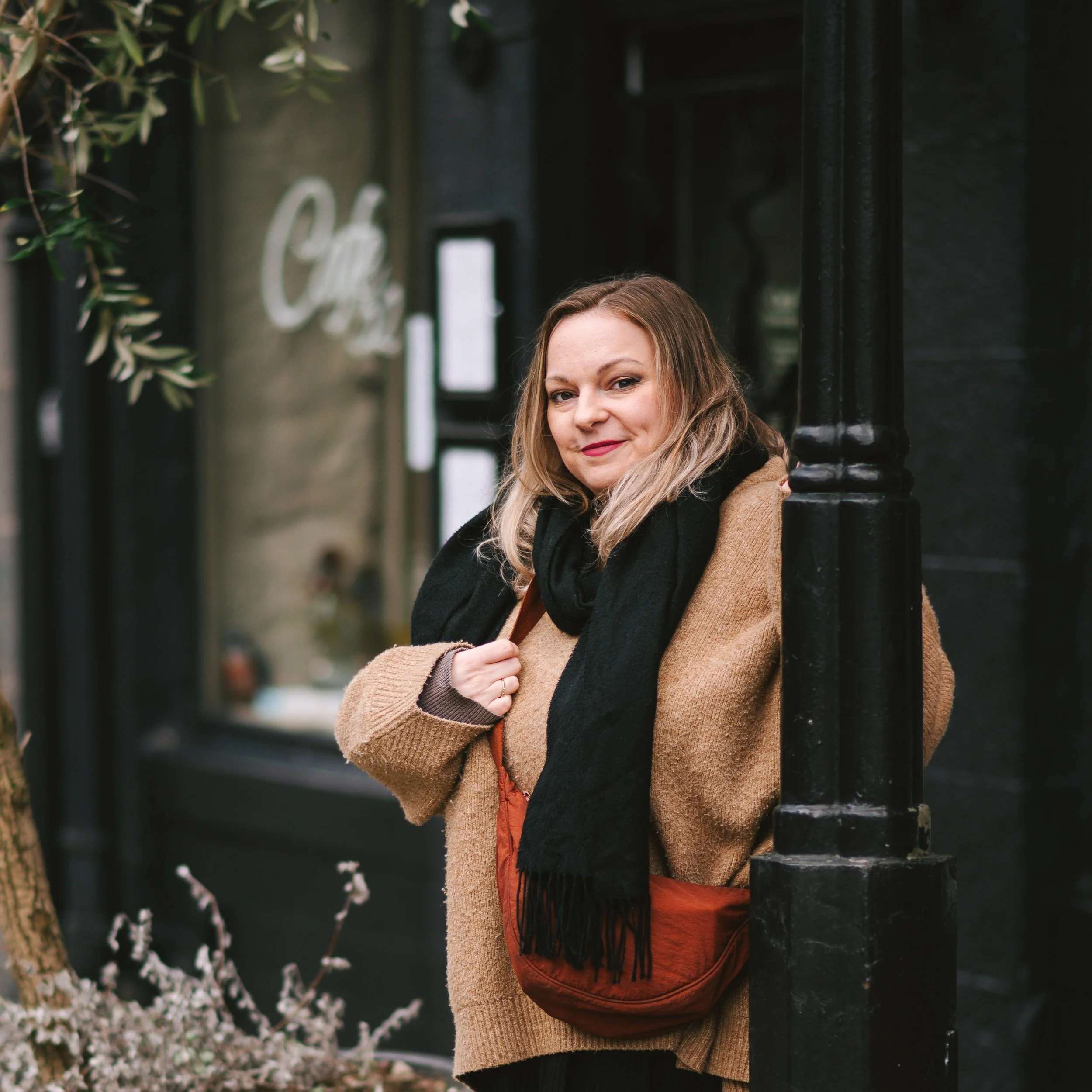 A woman with shoulder-length blonde hair and a smile, wearing a tan sweater, black scarf, and carrying a brown shoulder bag, standing outside near a black lamppost with a storefront in the background.
