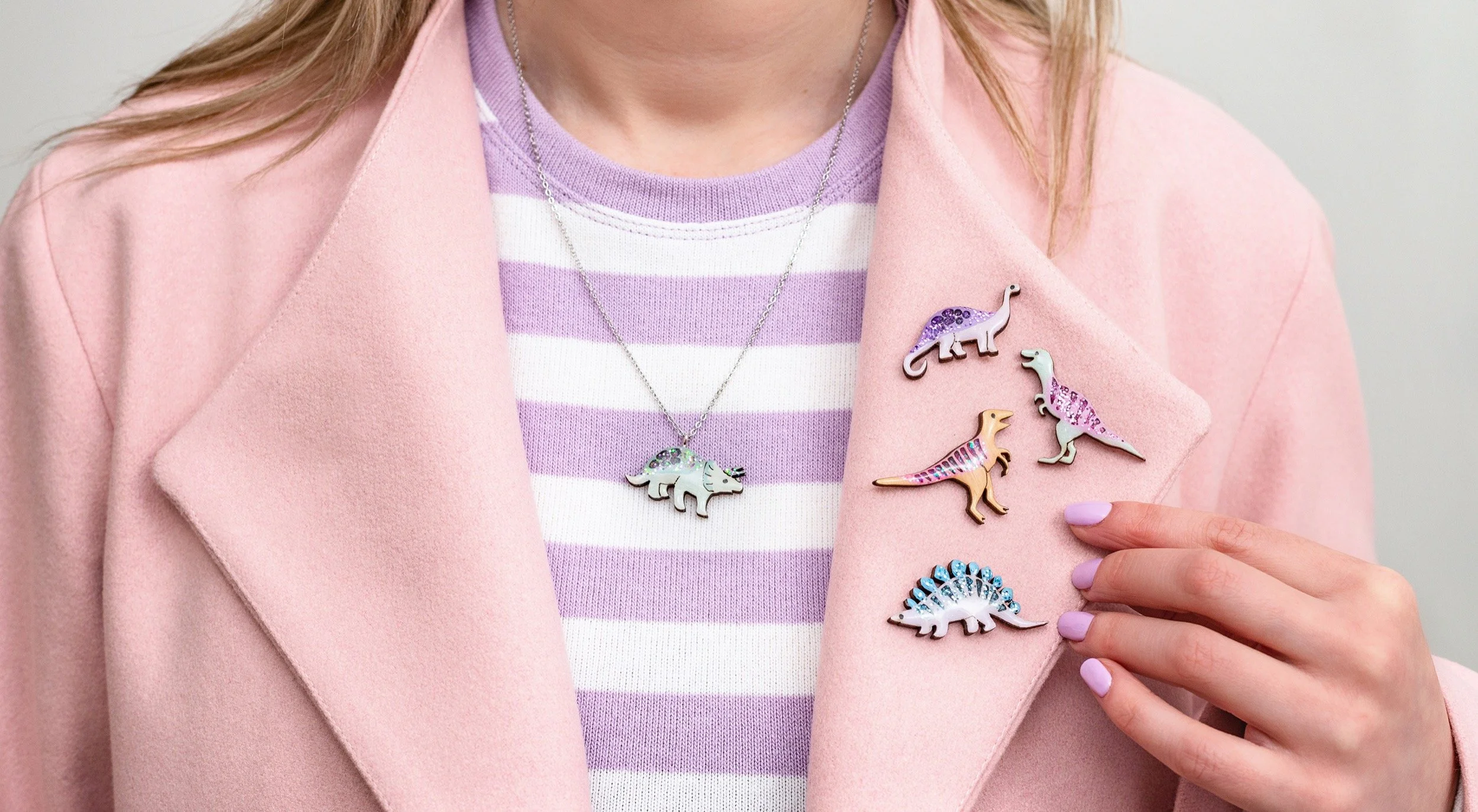 Woman in pink blazer wearing a purple and white striped shirt, displaying a dinosaur necklace and five dinosaur-shaped pins on her blazer.