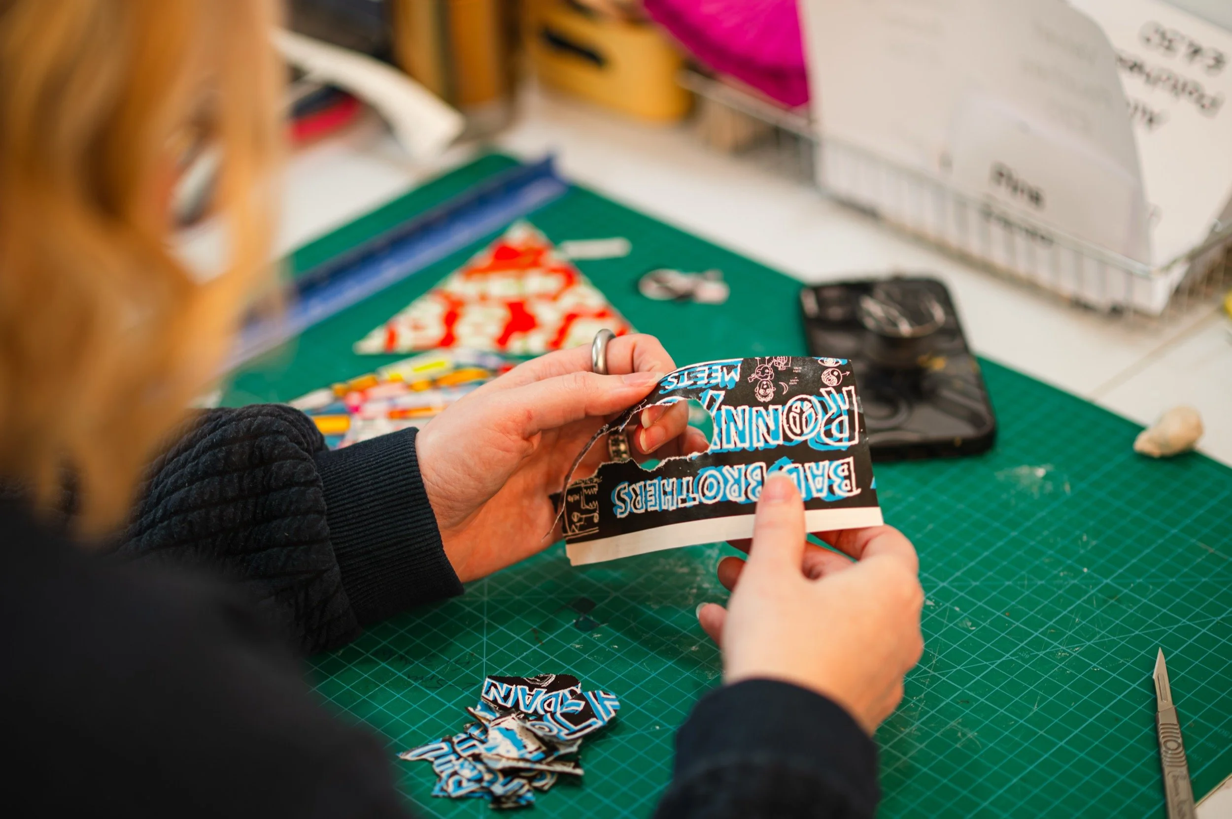 Person cutting a magazine with a pair of scissors at a craft workspace with various crafting supplies.