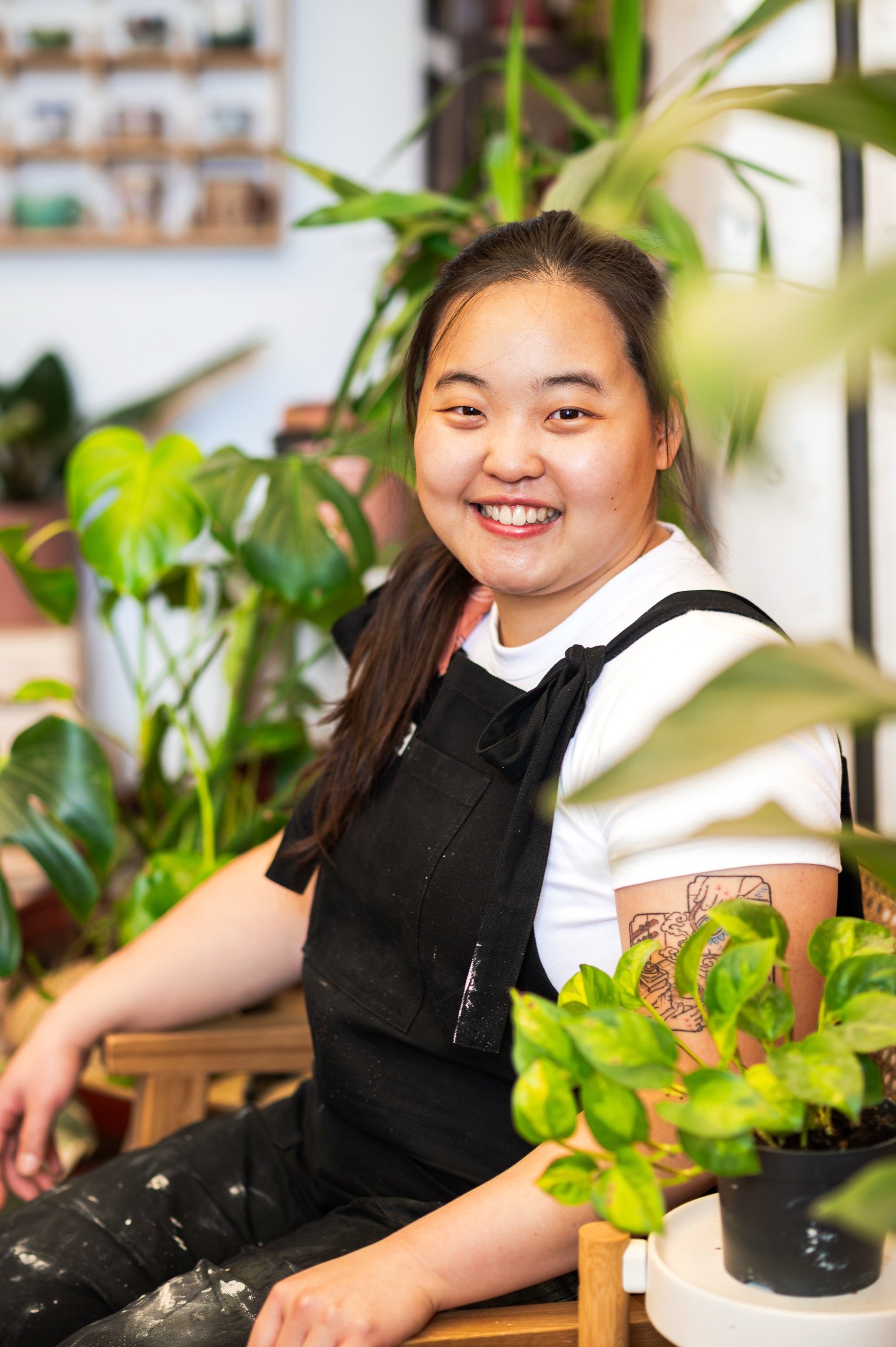 A young woman smiling while sitting among green houseplants in an indoor garden or plant shop.