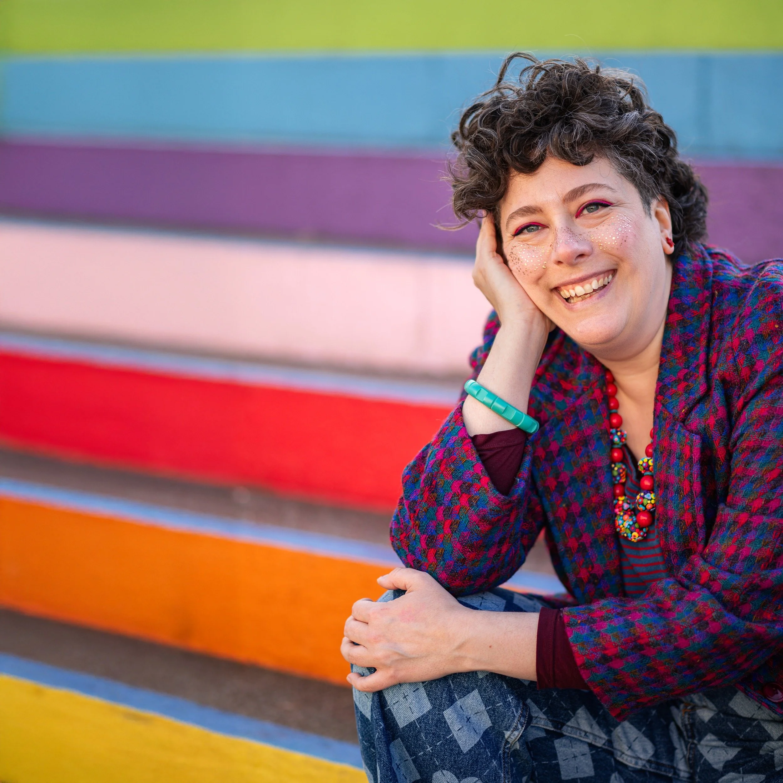 A smiling person with short curly hair and glitter makeup, sitting on colorful steps, wearing a multicolored patterned jacket, striped shirt, and colourful jewelry.