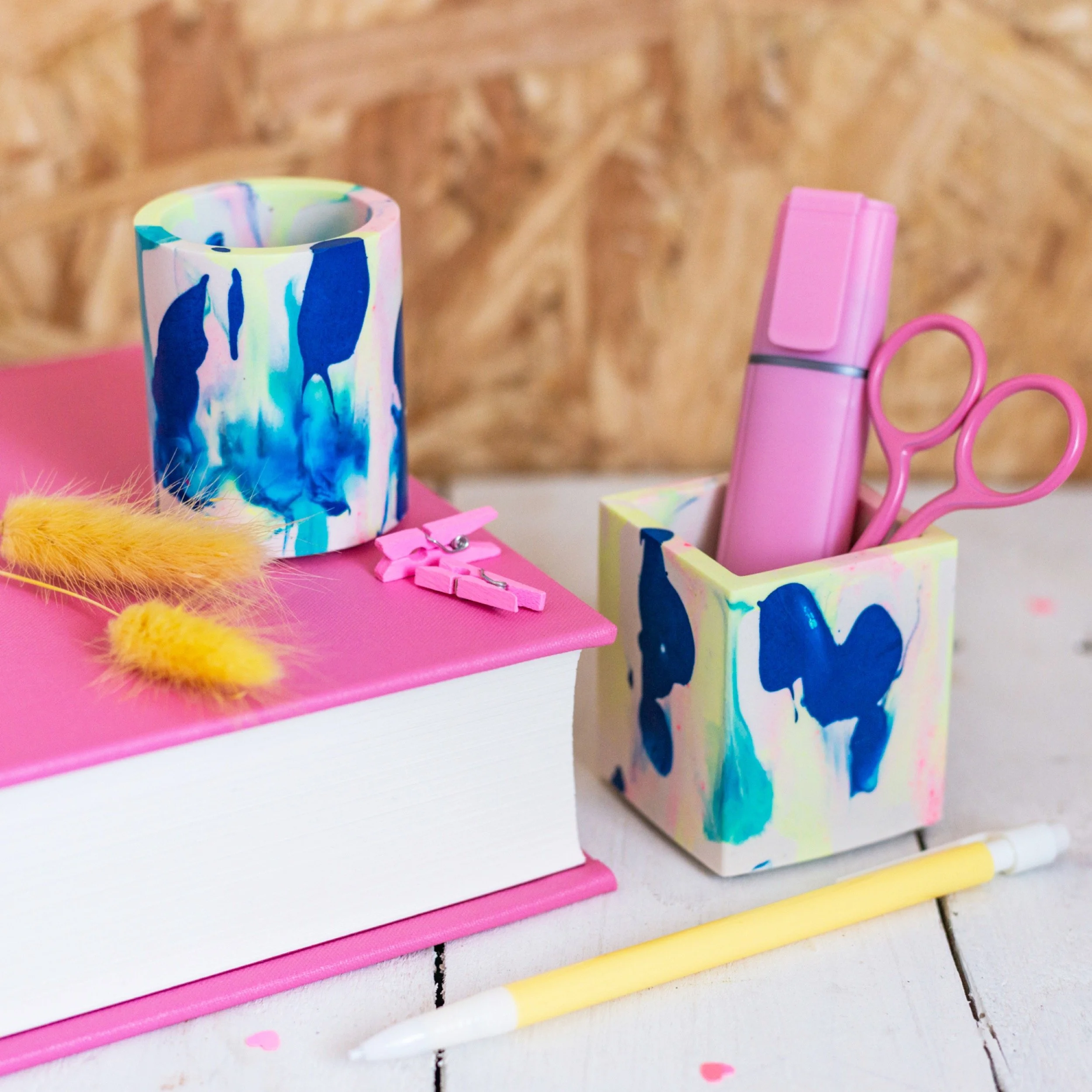 Desk organiser with pastel pink and blue art supplies, a pink pen, scissors, mini pegs, and a colourful tumbler on a pink book.