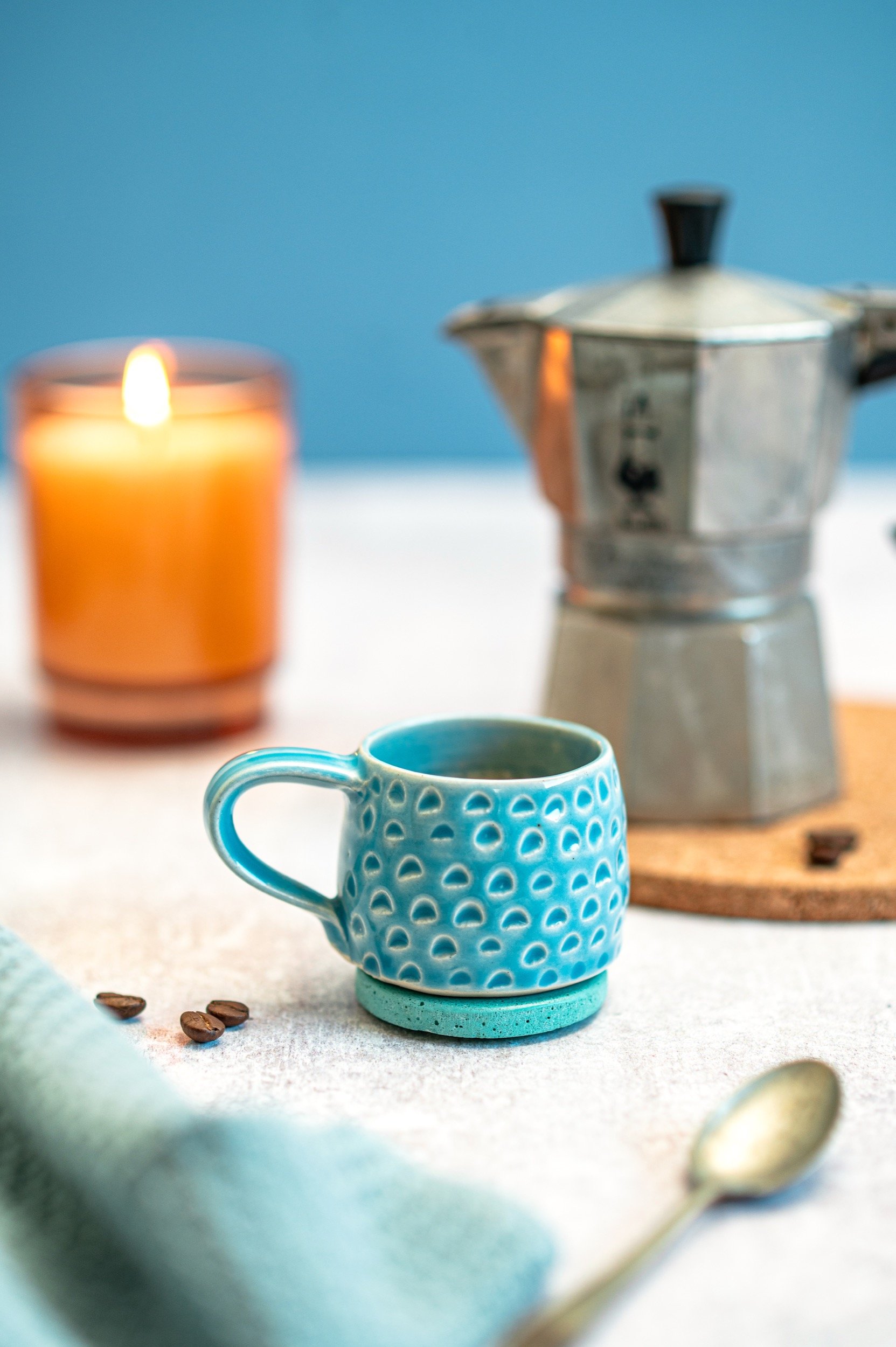 A blue textured ceramic coffee mug on a white surface with coffee beans nearby, a lit candle, a moka pot, and a spoon in the background.