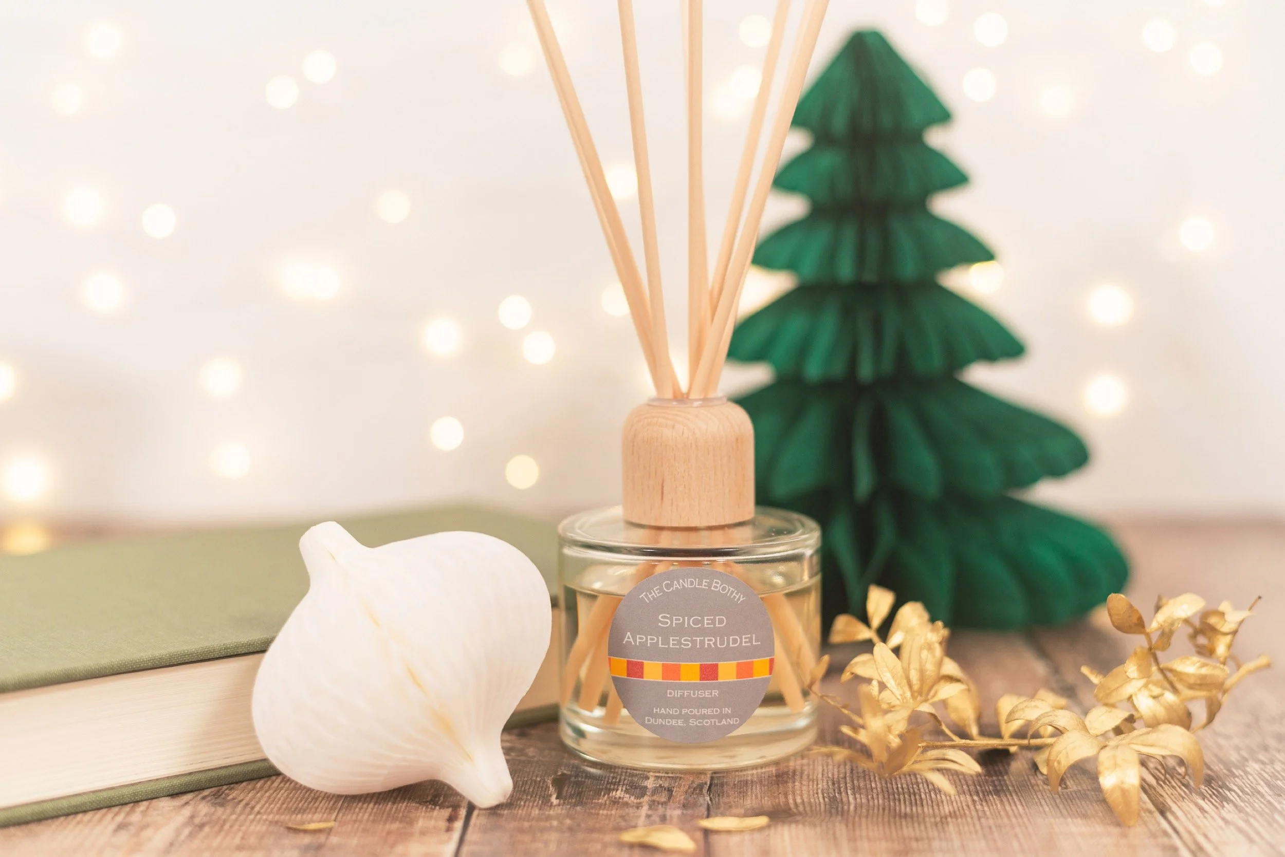 A glass candle diffuser labeled 'Spiced Applestrudel' with wooden reeds inside, a white shell, a green Christmas tree decoration, a closed book, and gold-colored leaves on a wooden surface, with blurred string lights in the background.