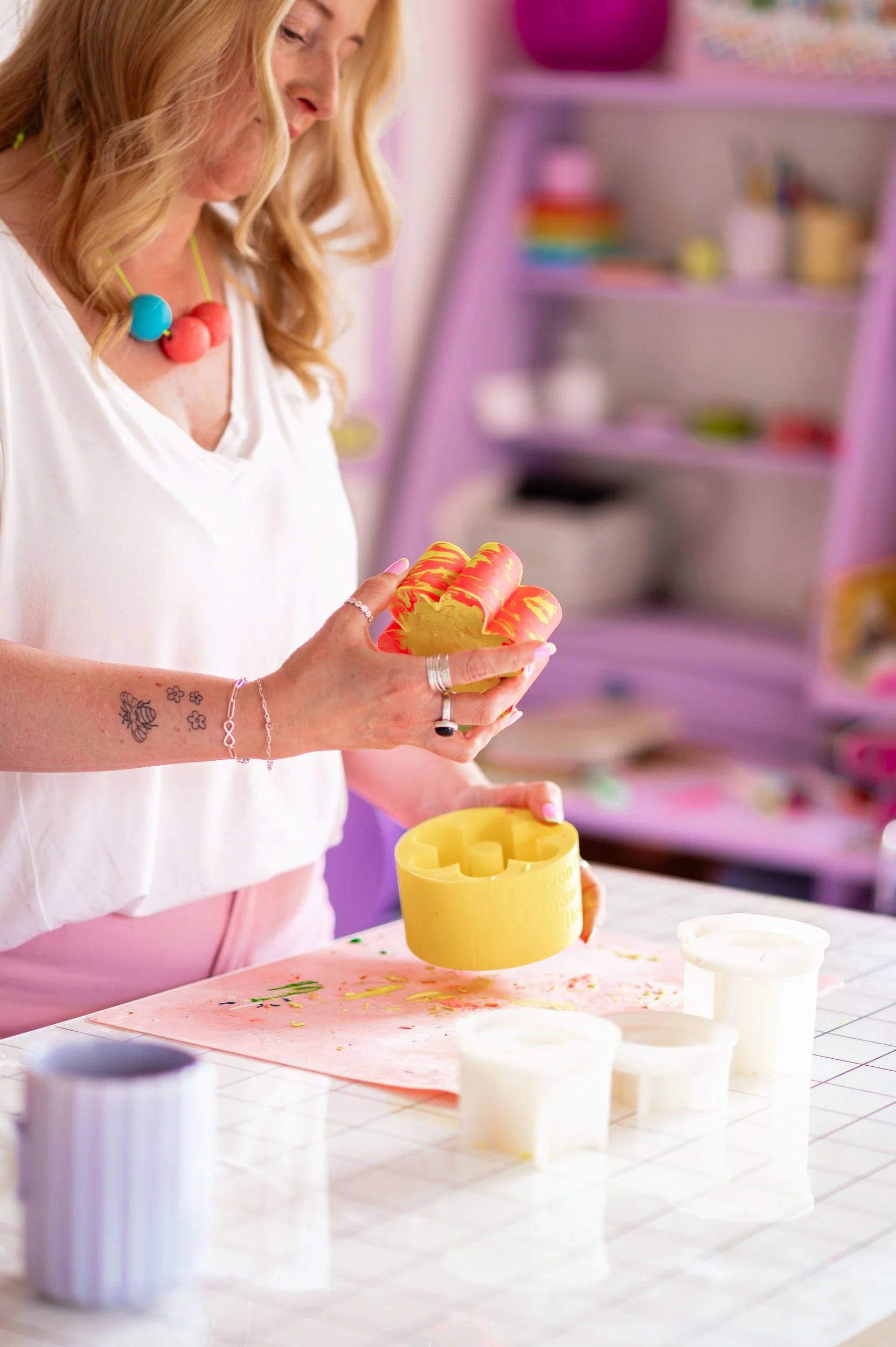 A woman with blonde hair, wearing a white shirt and colorful necklace, is holding a yellow and pink patterned jesmonite piece and silicone mold. She is in a studio with purple shelves in the background and various crafting supplies on the table.