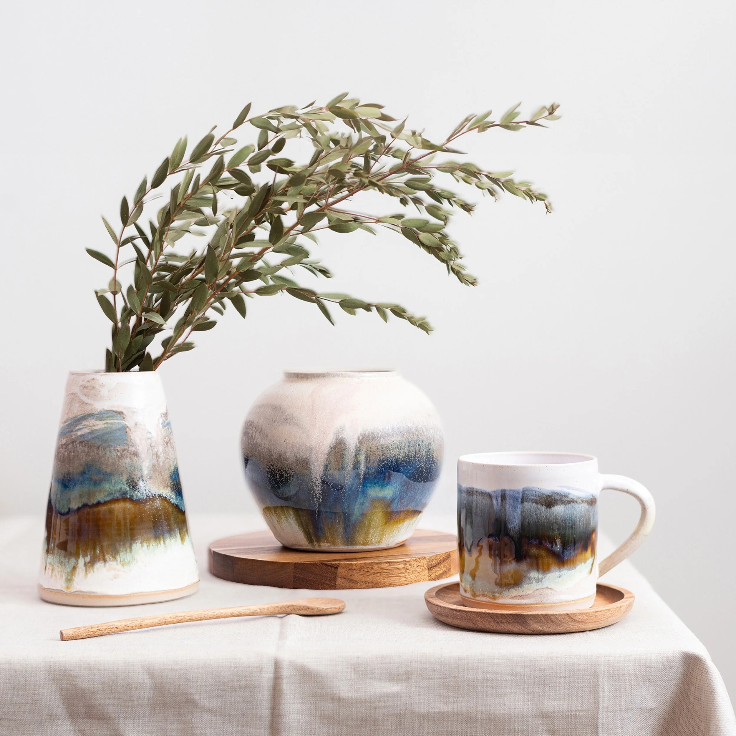 Decorative ceramic vases and mug with abstract watercolor design, placed on a table with linen tablecloth, holding greenery and accompanied by wooden coasters and a small wooden utensil.