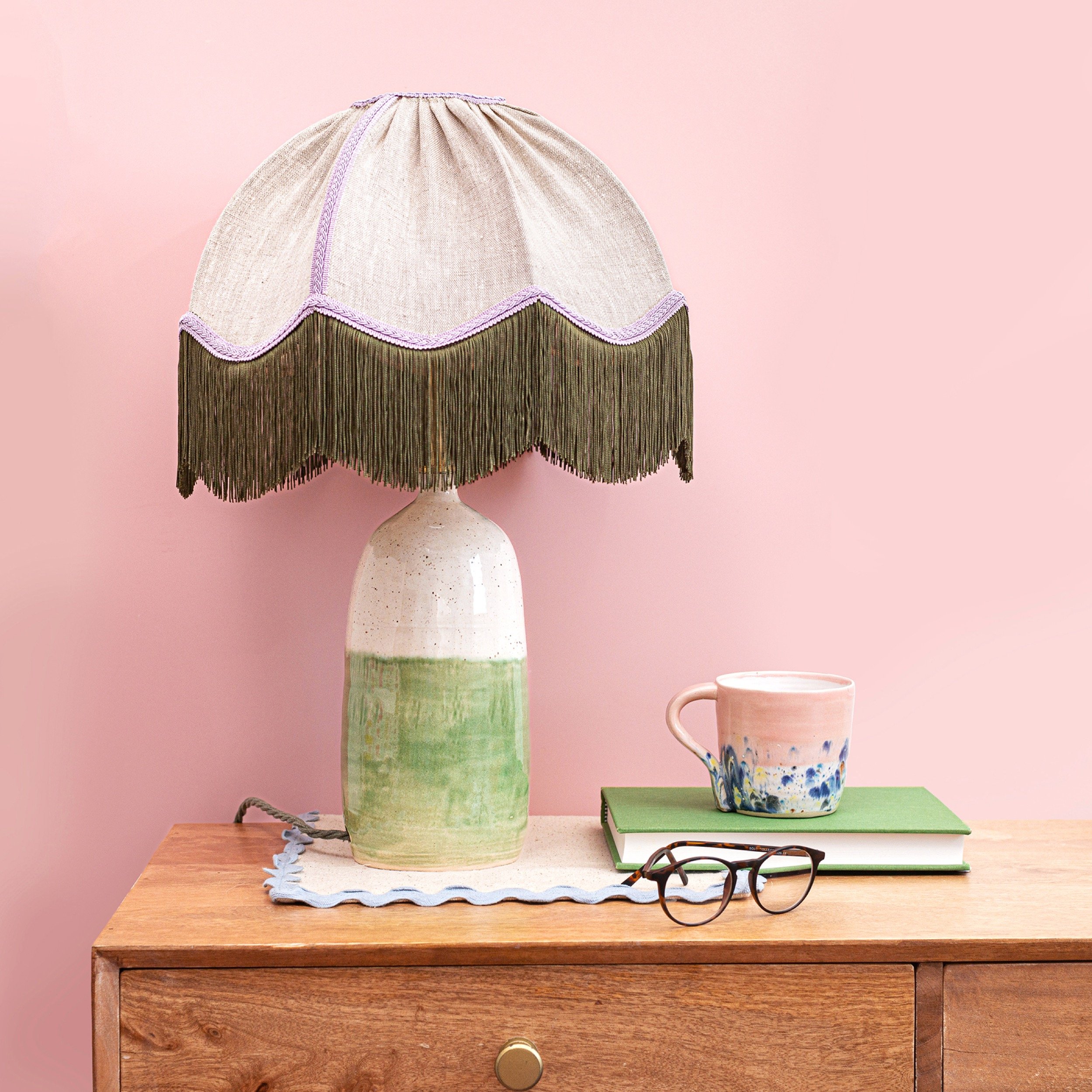 A wooden dresser with a tablecloth, a ceramic table lamp with a beige shade and fringe, a pink and blue coffee mug, a closed green notebook, a pair of black-rimmed glasses, all against a pink wall.
