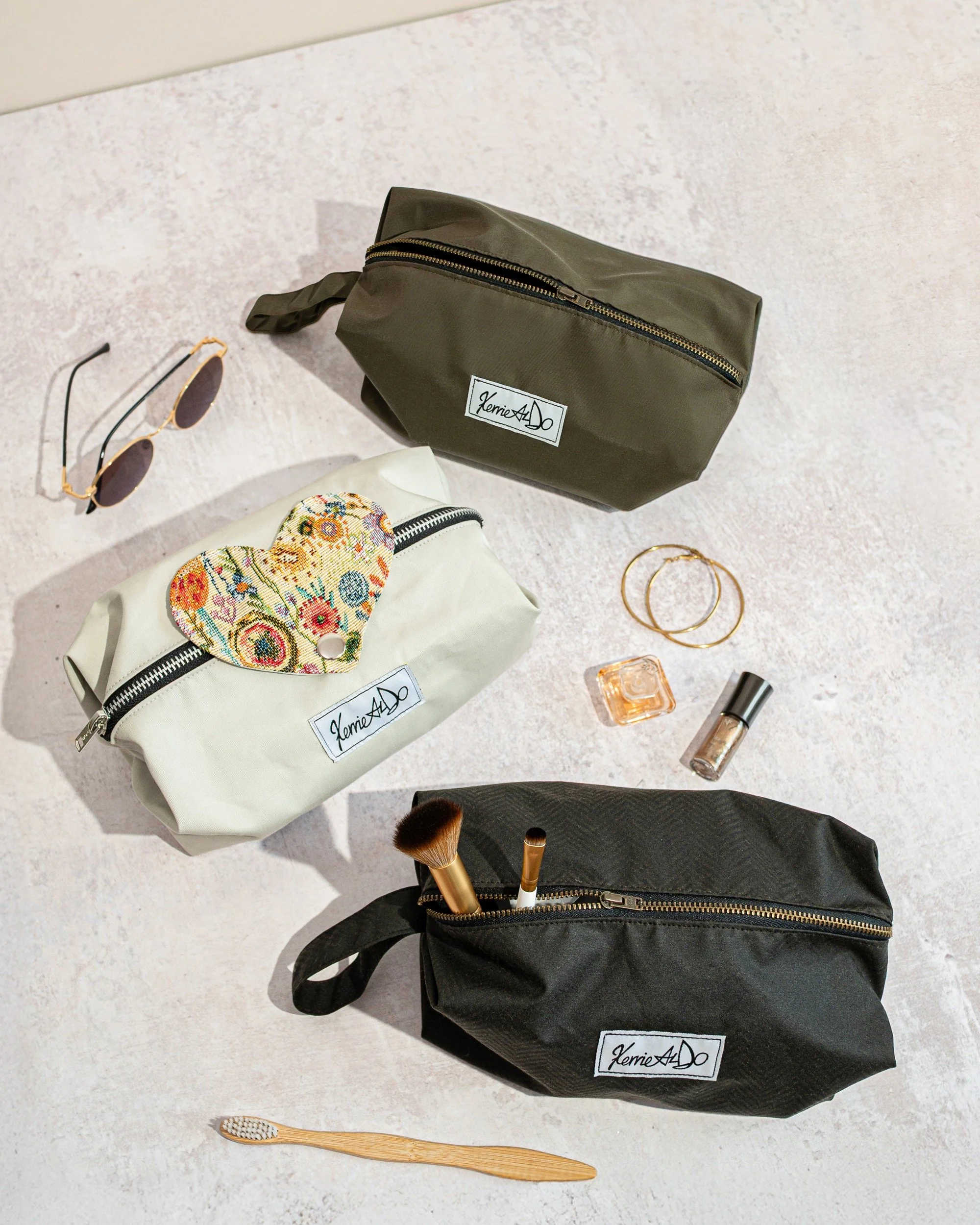 Three cosmetic bags on a light-coloured surface with various items including sunglasses, gold earrings, a small perfume bottle, glitter nail polish, a makeup brush, and a bamboo toothbrush.
