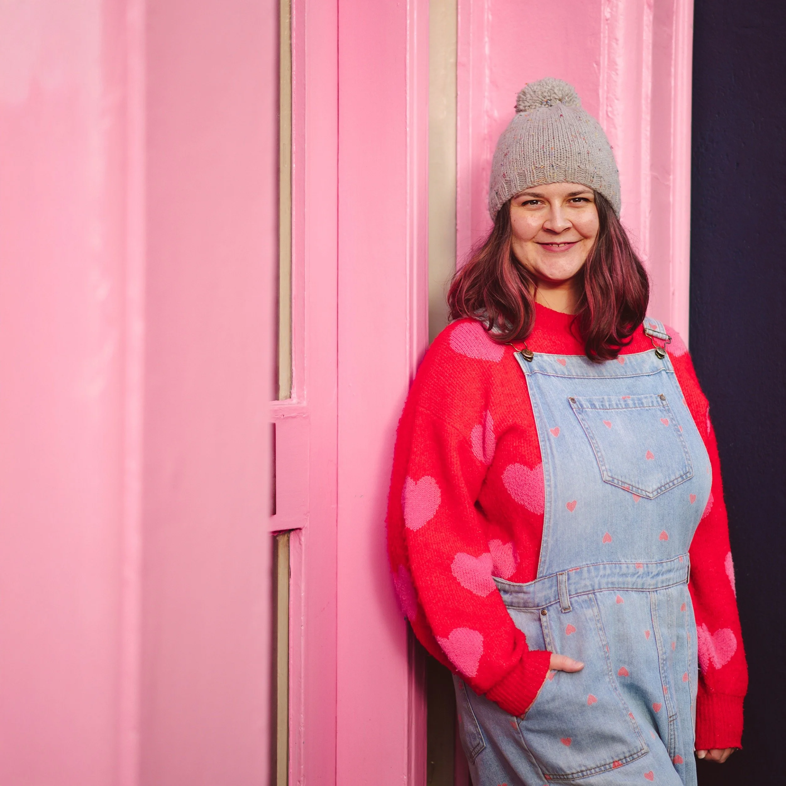 Hello! Jilly smiles, looking at the camera. She stands in front of a pink shop.