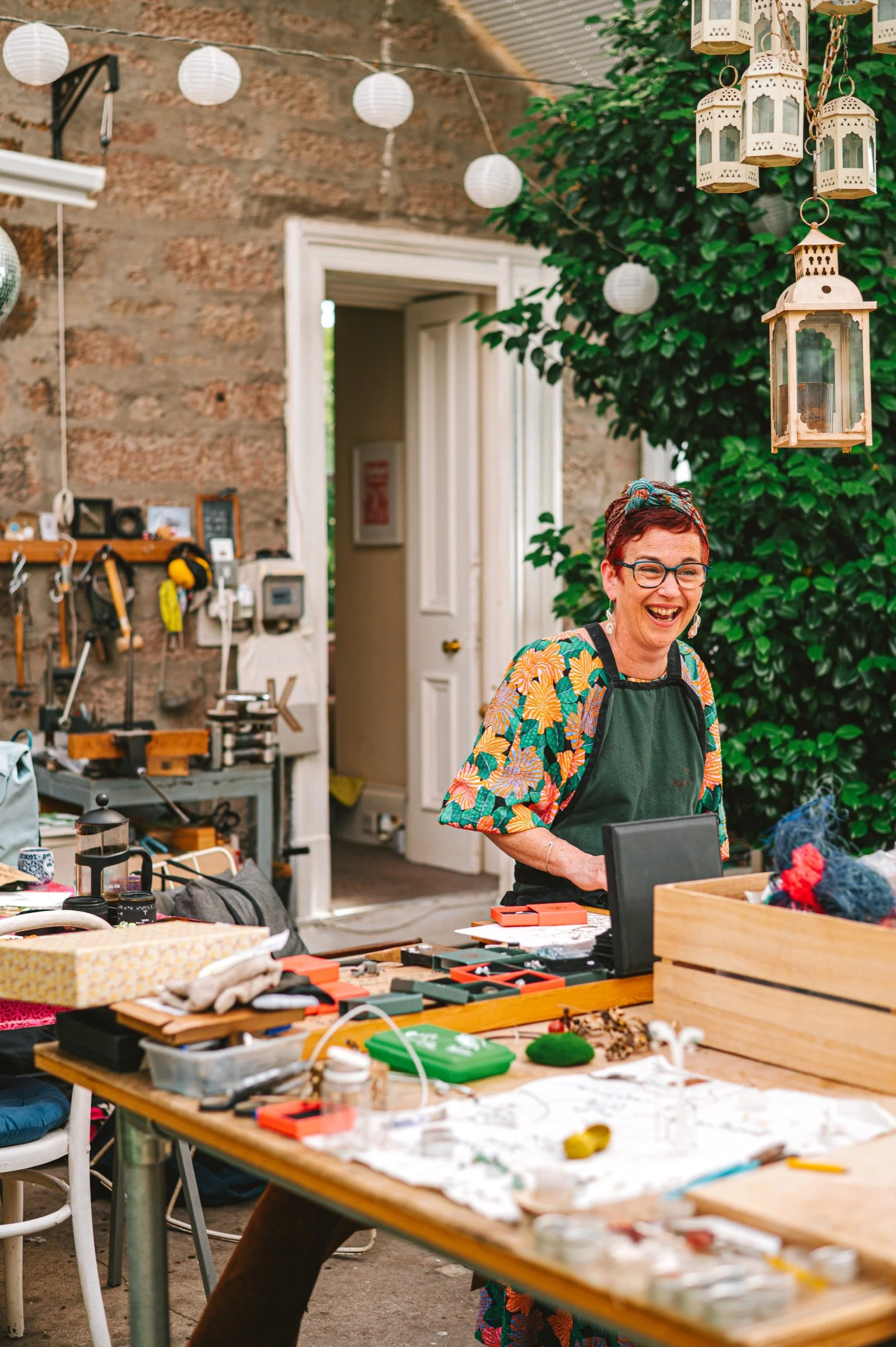 A woman with short red hair, glasses, and a colorful floral shirt laughing indoors. She is wearing a dark apron and standing behind a large wooden table cluttered with various craft supplies, jewelry pieces, and tools. The background includes a brick