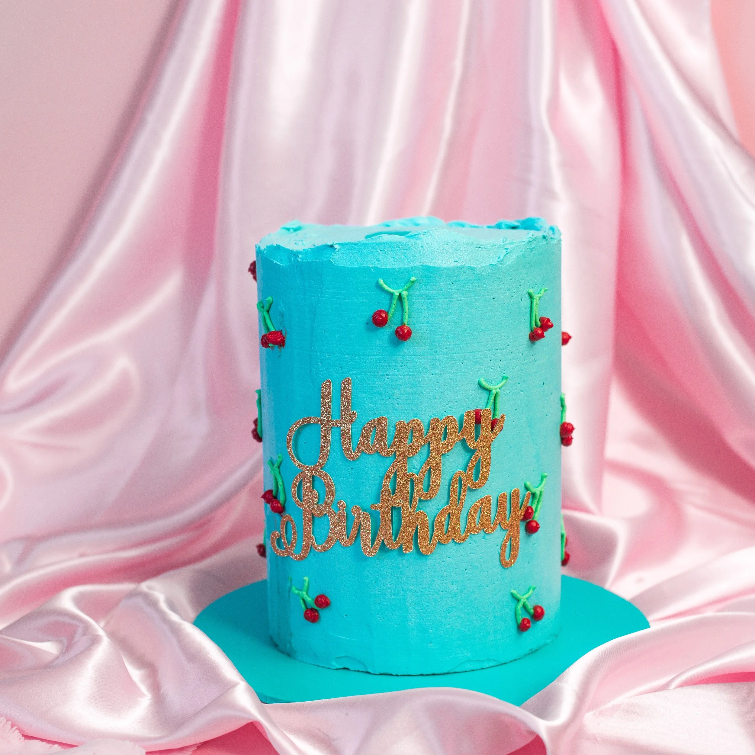 A tall, cylindrical birthday cake covered in blue frosting with red cherry decorations and a gold 'Happy Birthday' message, placed on a blue cake board with pink satin fabric in the background.