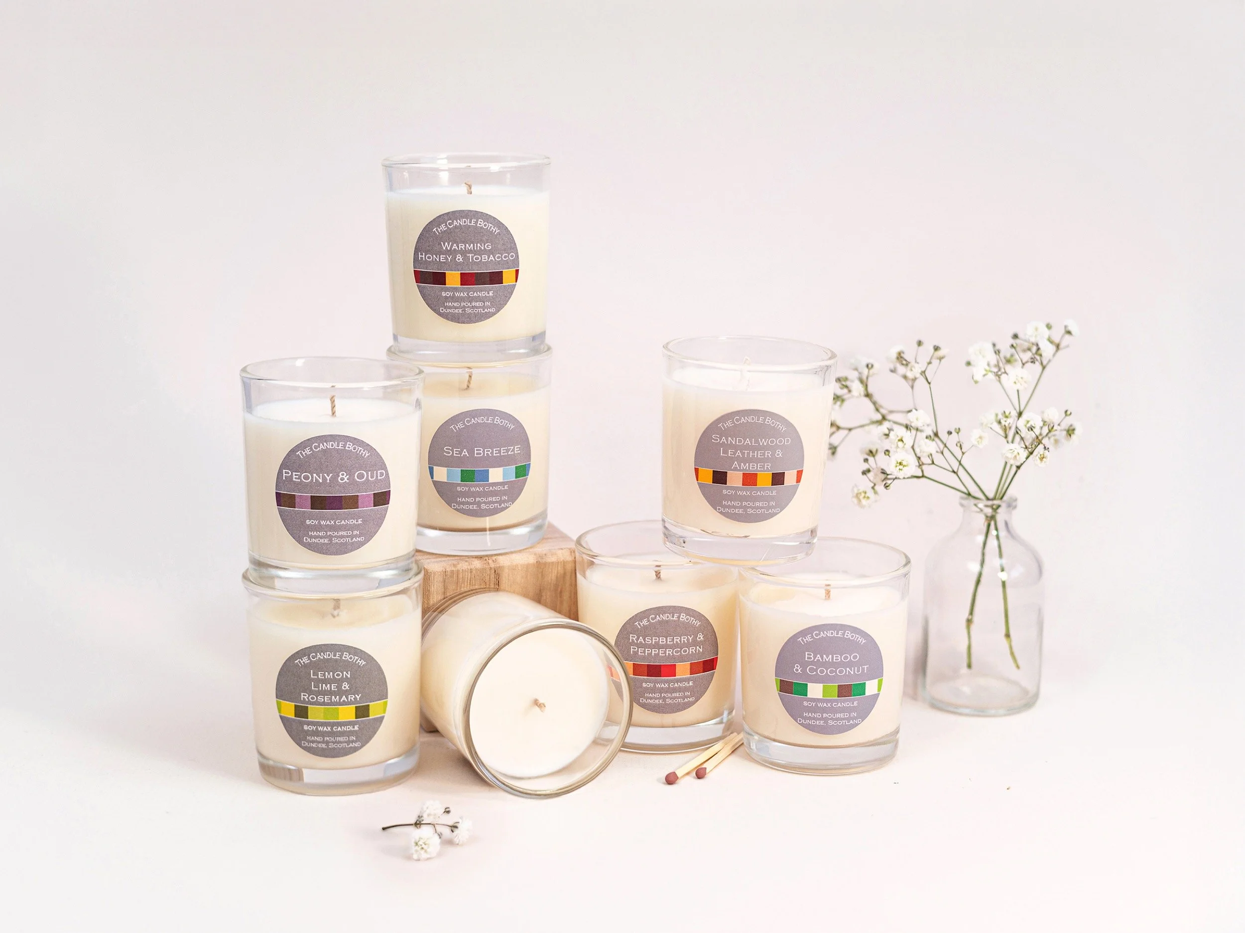 A collection of seven white candles in clear glass jars with colorful labels, arranged on a white surface with a small glass vase of white flowers to the right.