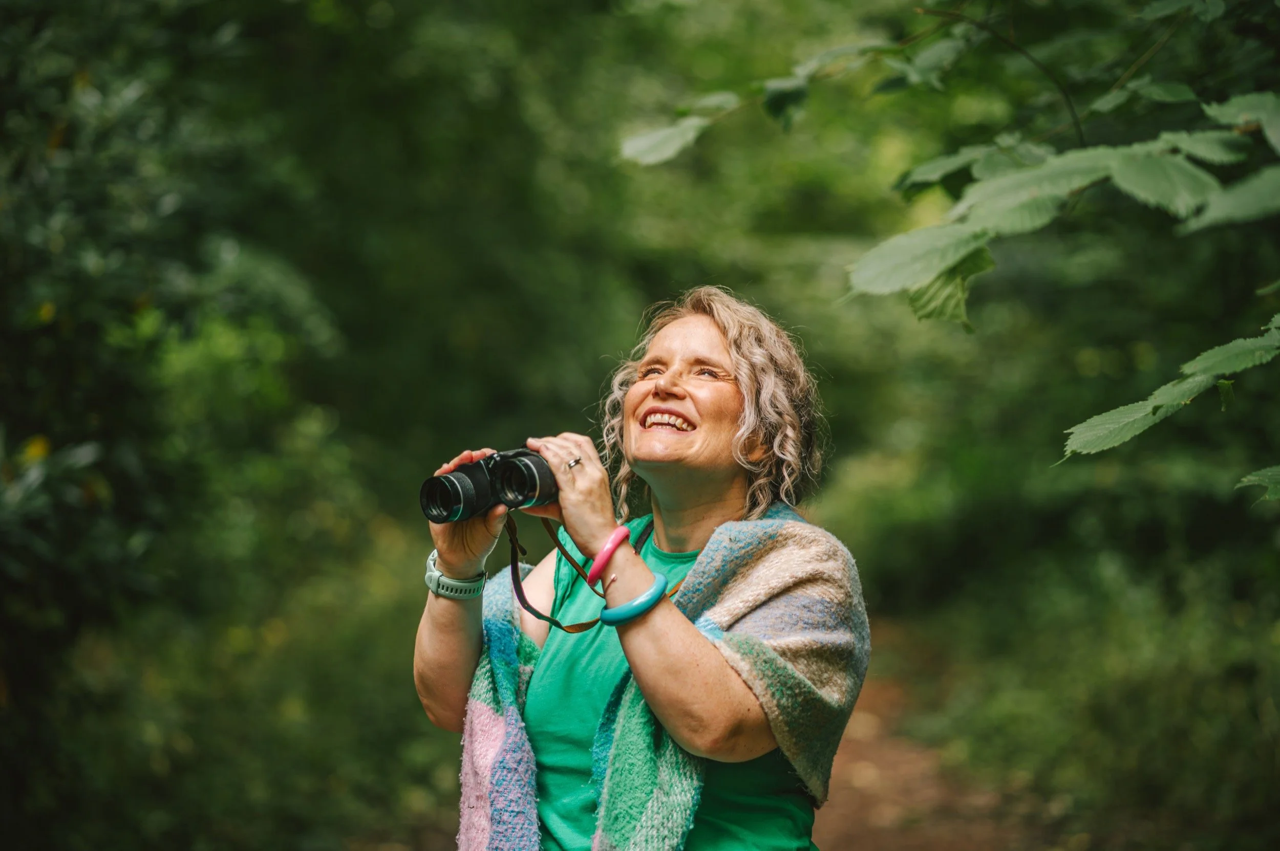 A woman with curly hair holding binoculars in a lush green forest, smiling and looking upwards.