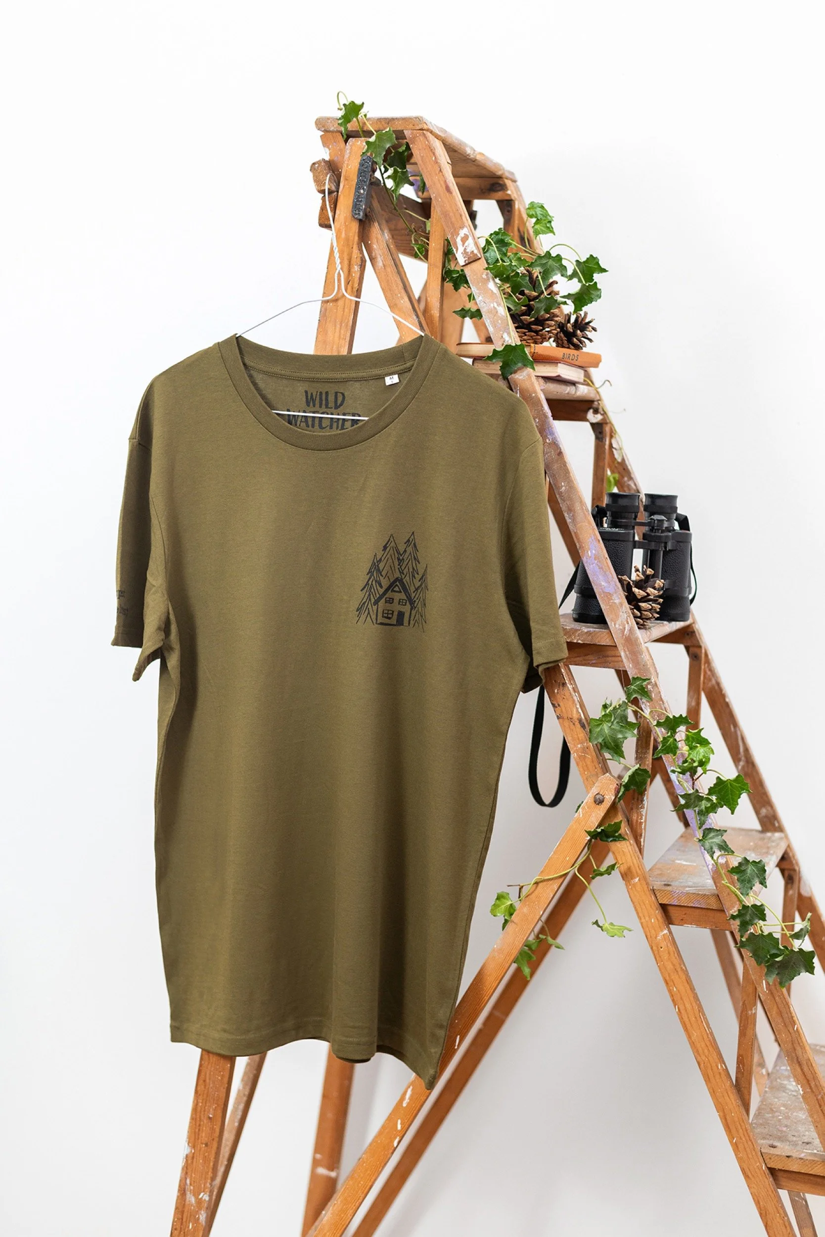 An olive green T-shirt with a tree and house graphic, hanging on a hanger in front of a wooden ladder decorated with books, pinecones, binoculars, and green ivy plants, against a white wall.
