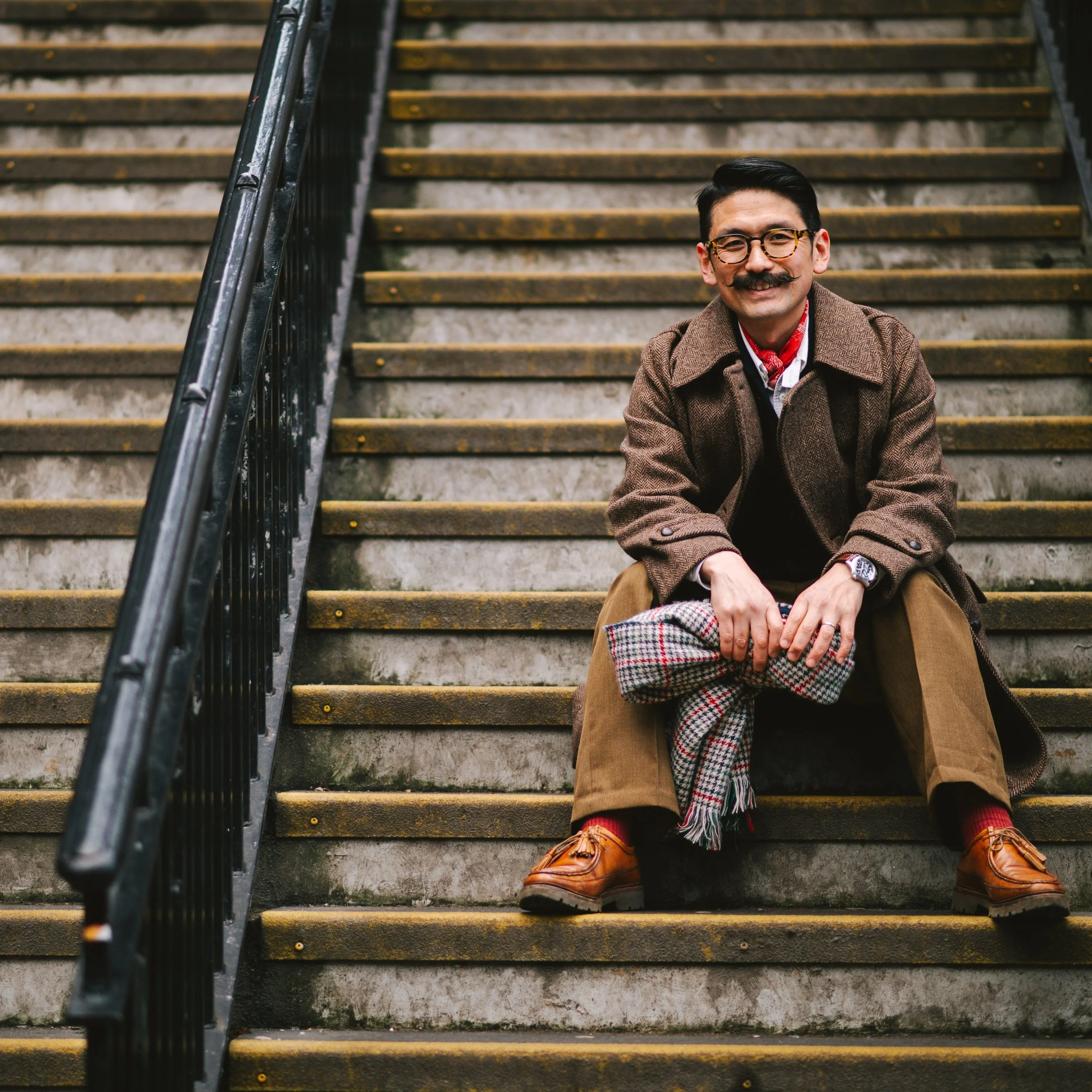 A smiling man with glasses and a mustache sitting on outdoor stairs, holding a scarf, wearing a brown coat, tan trousers, and brown shoes.