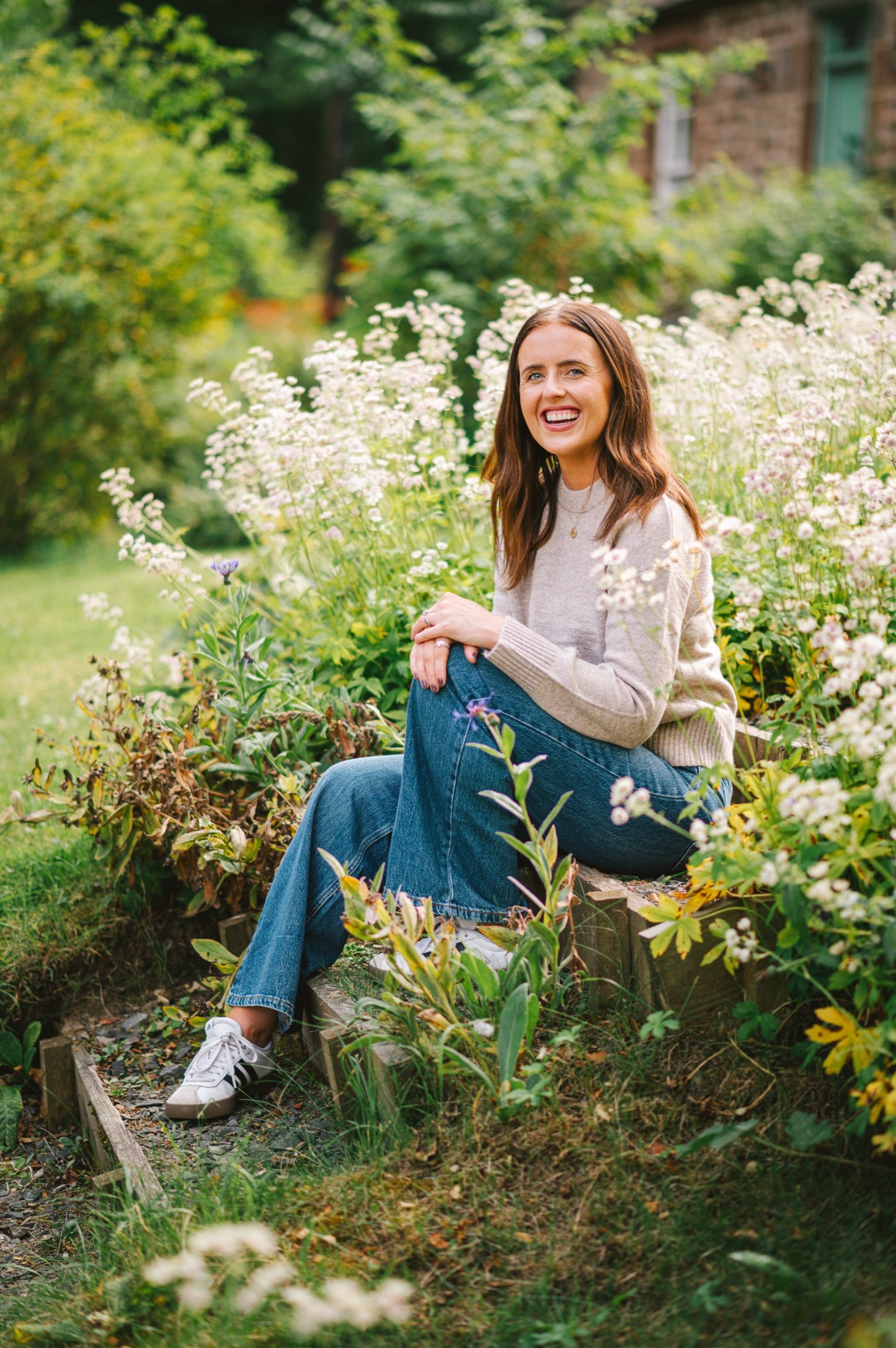 A woman sitting on a small wooden step among flowering plants in a garden, smiling and looking at the camera.