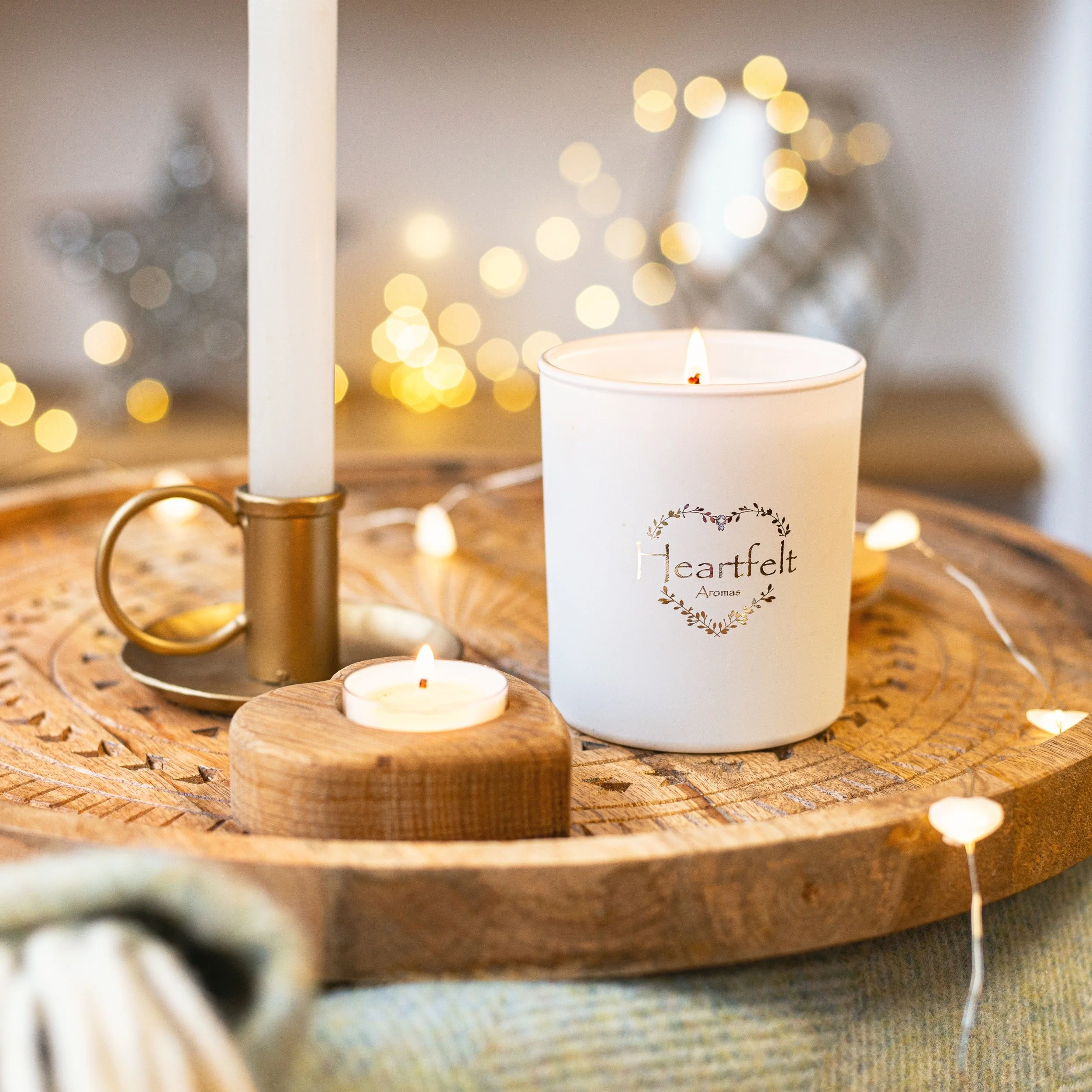 A wooden tray with candles, including a large white candle labeled 'Heartfelt Aromas' and a smaller tealight candle, adorned with string lights and a decorative silver star in the background.