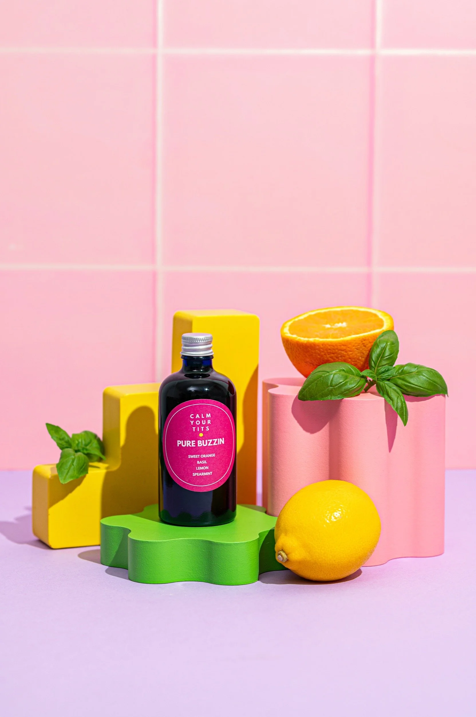 A pink background with colorful geometric blocks, a black bottle labeled 'Pure Buzzin,' a lemon, an orange half, and fresh basil leaves.