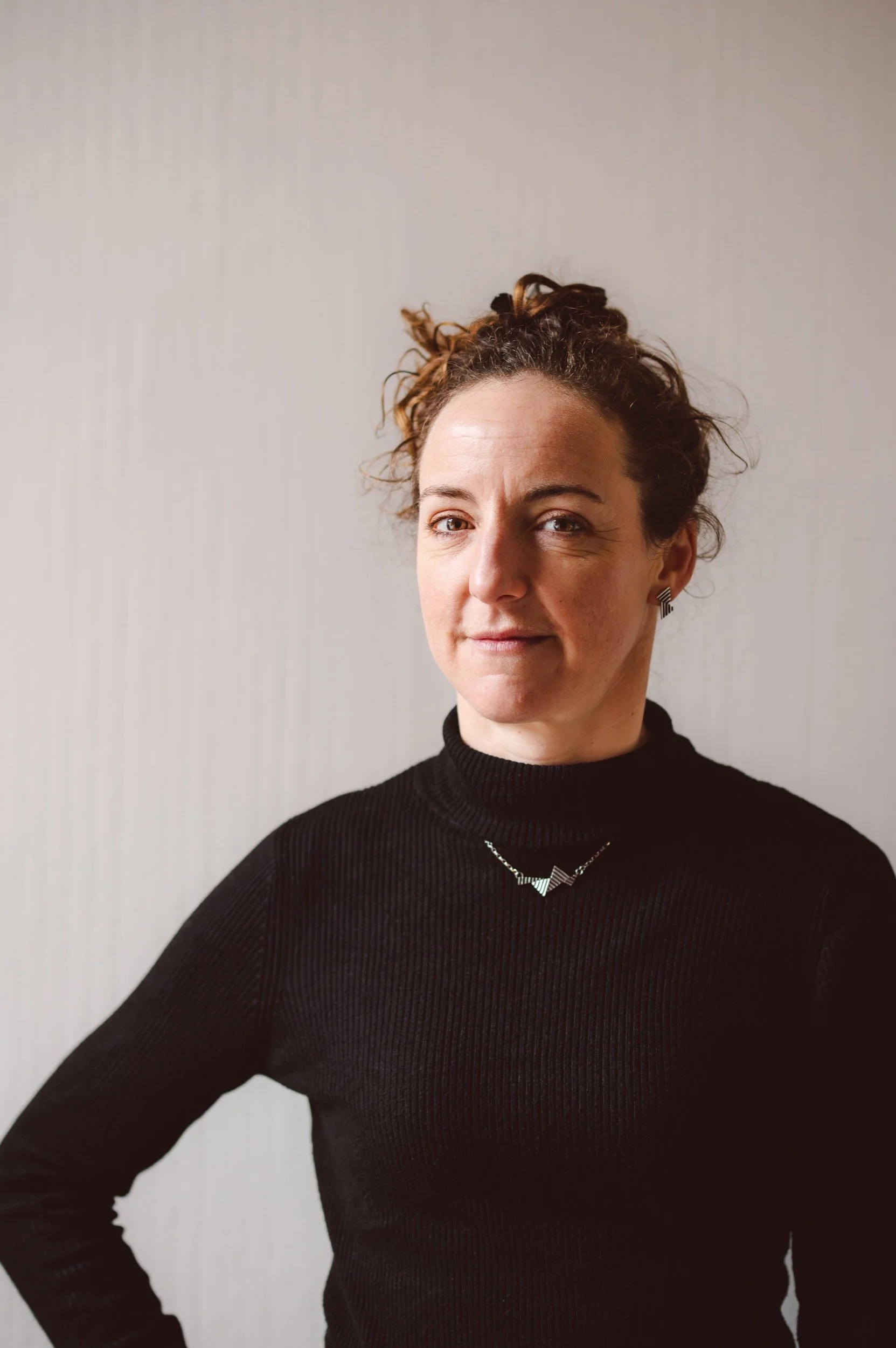 A woman with curly brown hair tied up, wearing a black turtleneck sweater, a necklace, and earrings, standing against a light-colored textured wall, looking confidently at the camera.