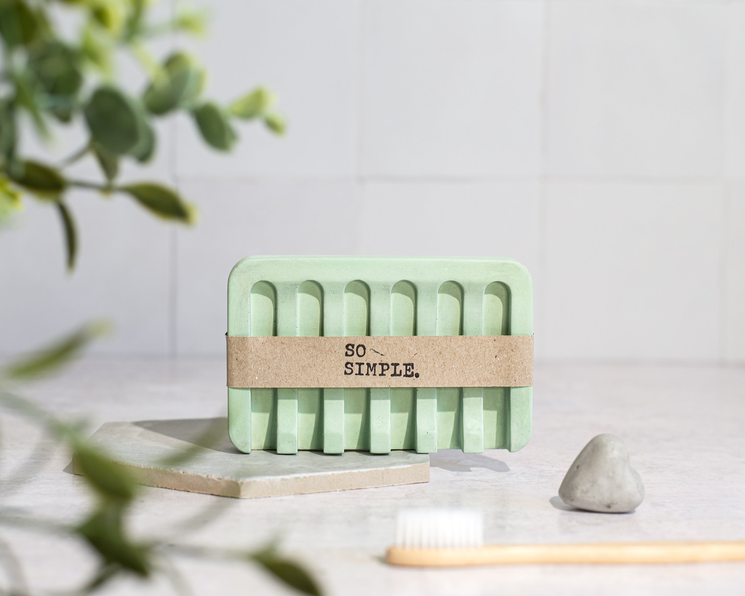 A green soap dish with a brown paper band that has the words 'SO SIMPLE.' printed on it, placed on a tiled surface in a minimalistic setting. A small white stone and a toothbrush are also visible.