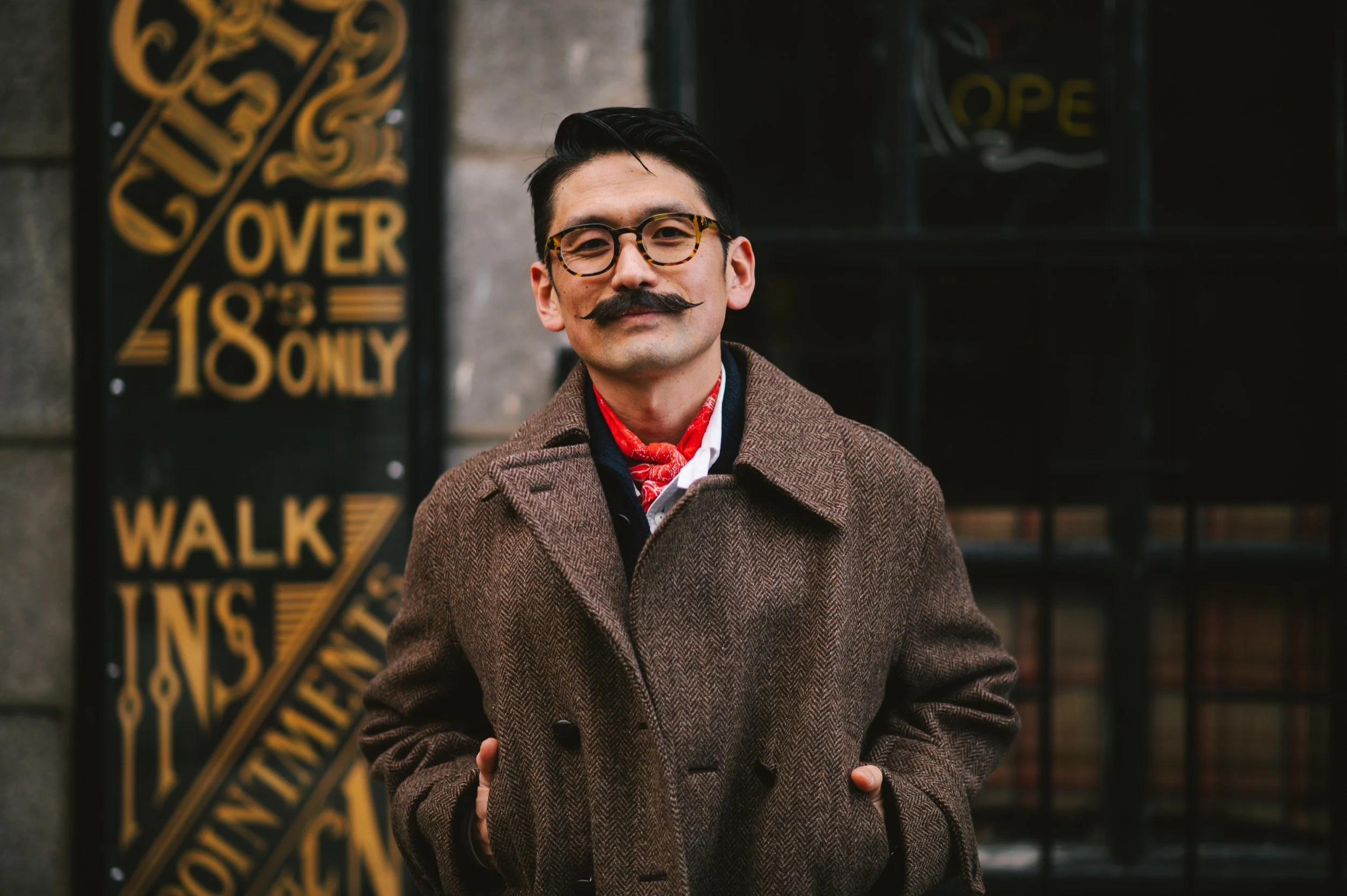 A man standing outdoors in front of a black sign with yellow lettering, wearing glasses, a brown coat, a red scarf, and a white shirt, with his hands in his coat pockets.