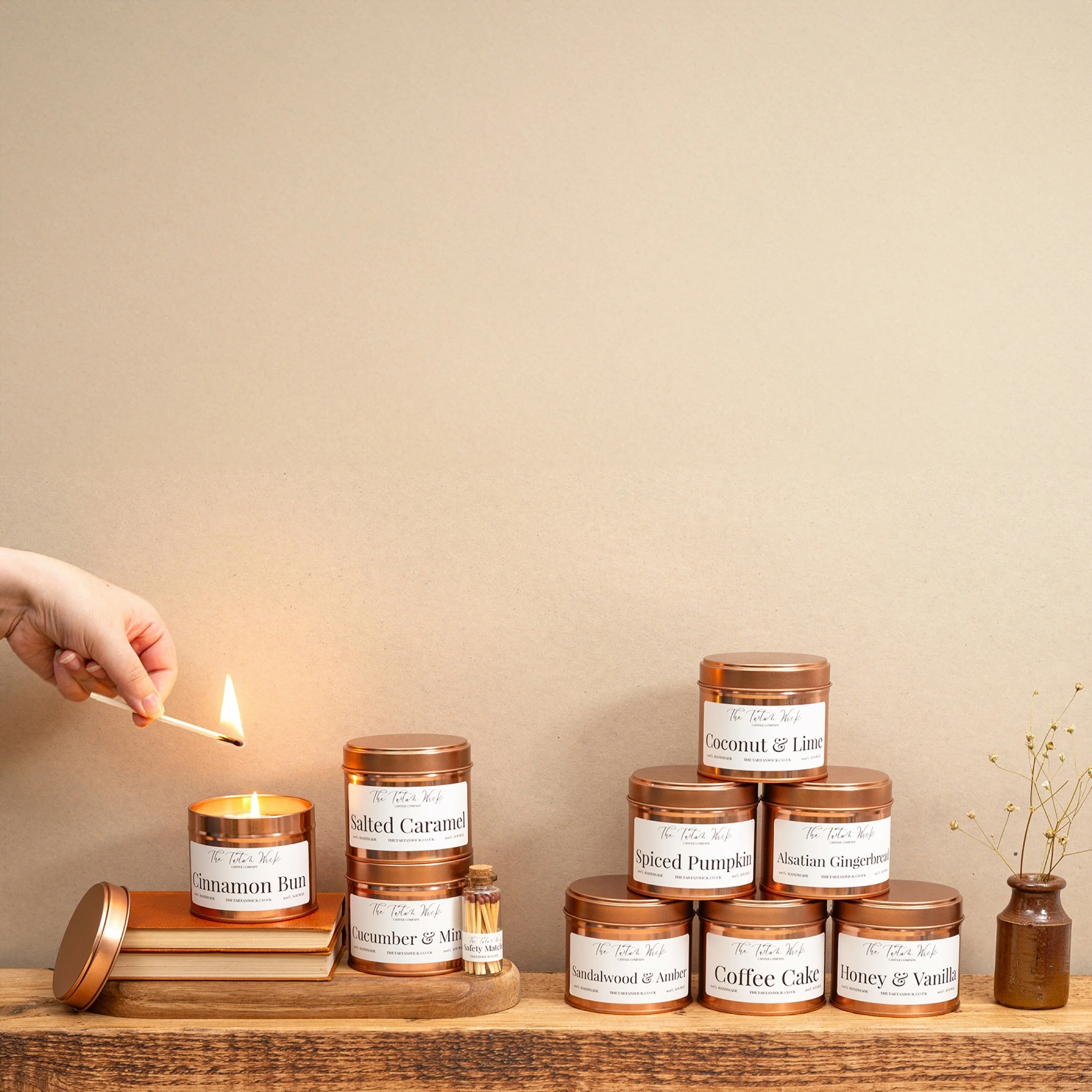 A collection of scented candles on a wooden surface, some labeled with flavours like Cinnamon Bun, Salted Caramel, Coconut & Lime, and others with various scents. A hand is lighting a candle with a matchstick. A small jar of matches is nearby.