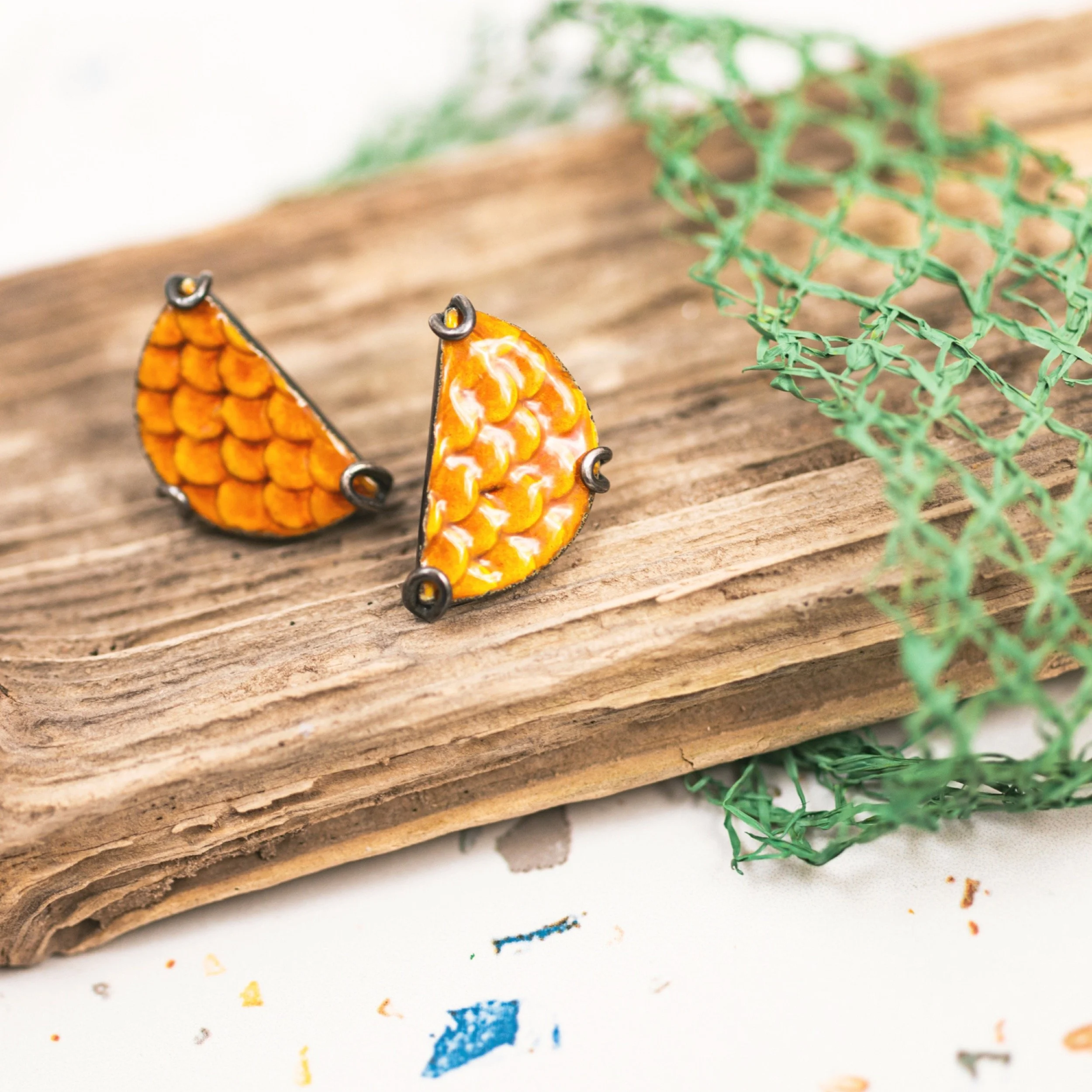 Pair of decorative earrings with orange and yellow scales design, placed on a piece of wood, with green decorative netting nearby.