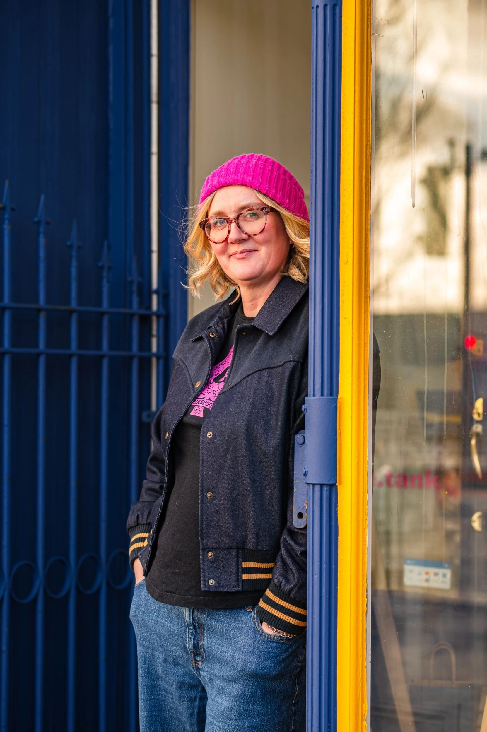 Woman with blond hair, glasses, pink beanie, and casual clothing standing outside near blue and yellow painted shop with a glass window.