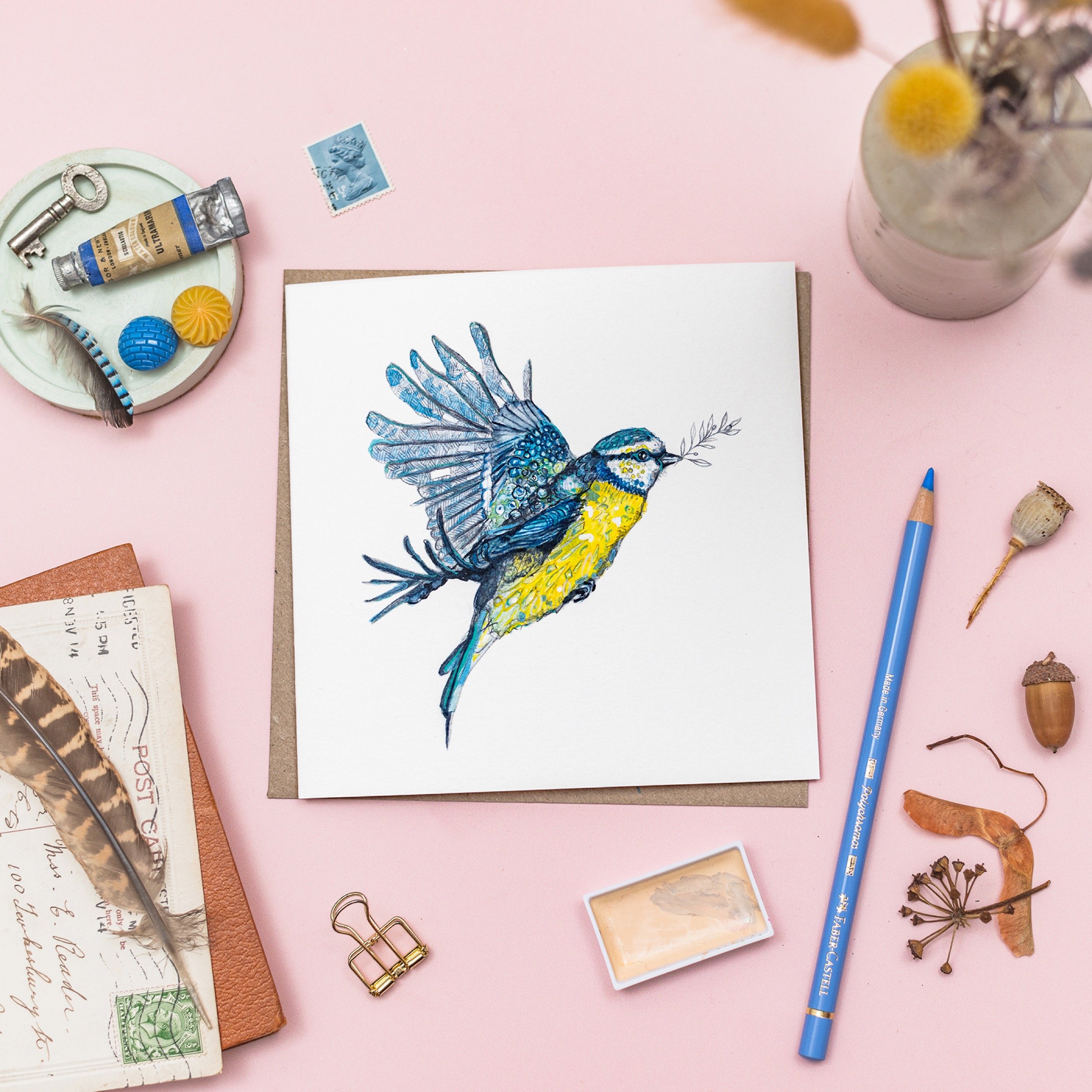 Illustrated card of a yellow and blue bird holding a sprig of leaves in its beak, surrounded by art materials on a pink background.