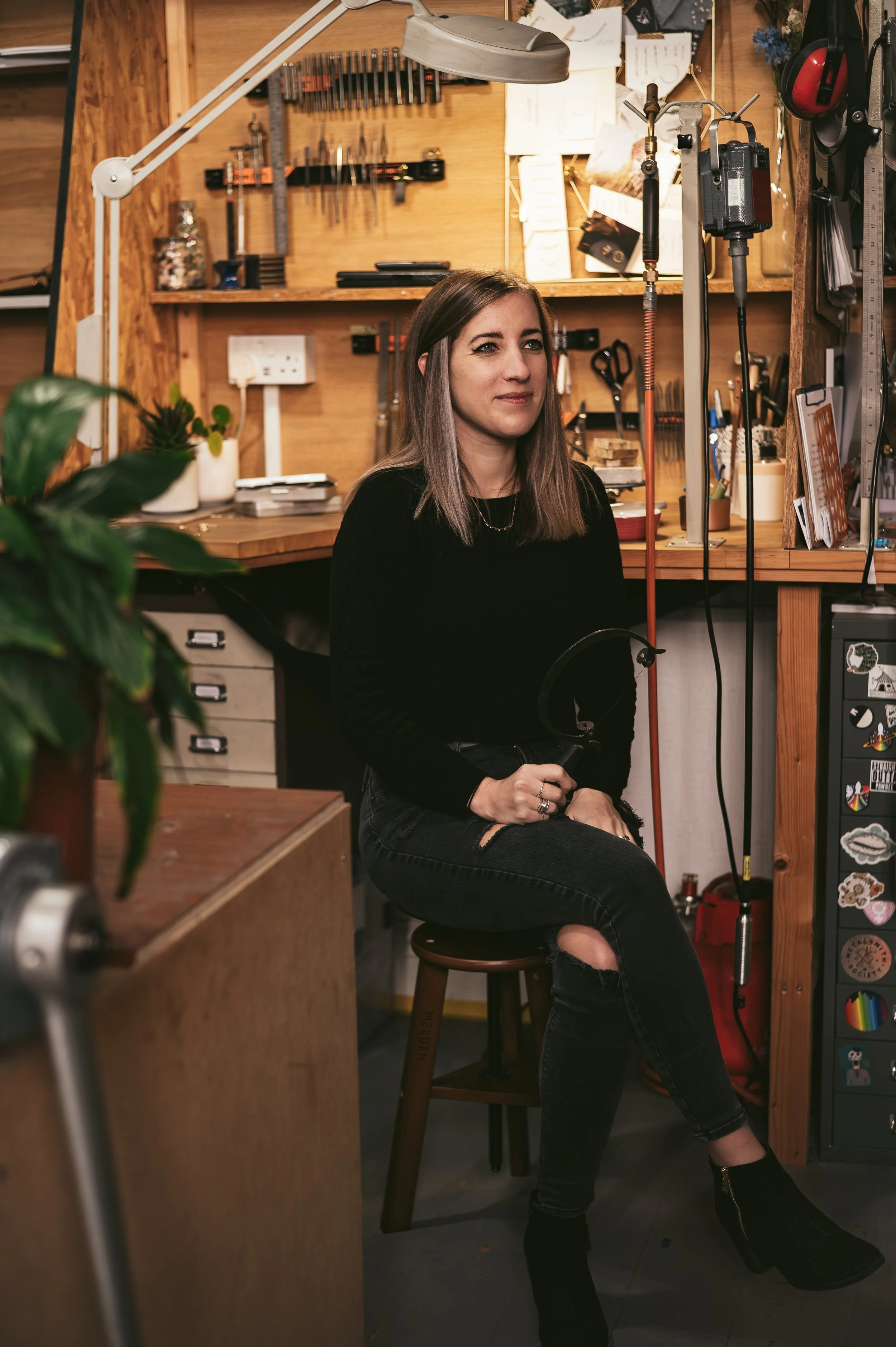 A woman with shoulder-length grayish hair sitting on a stool in a woodworking or craft workshop, surrounded by tools, wooden shelves, and equipment.