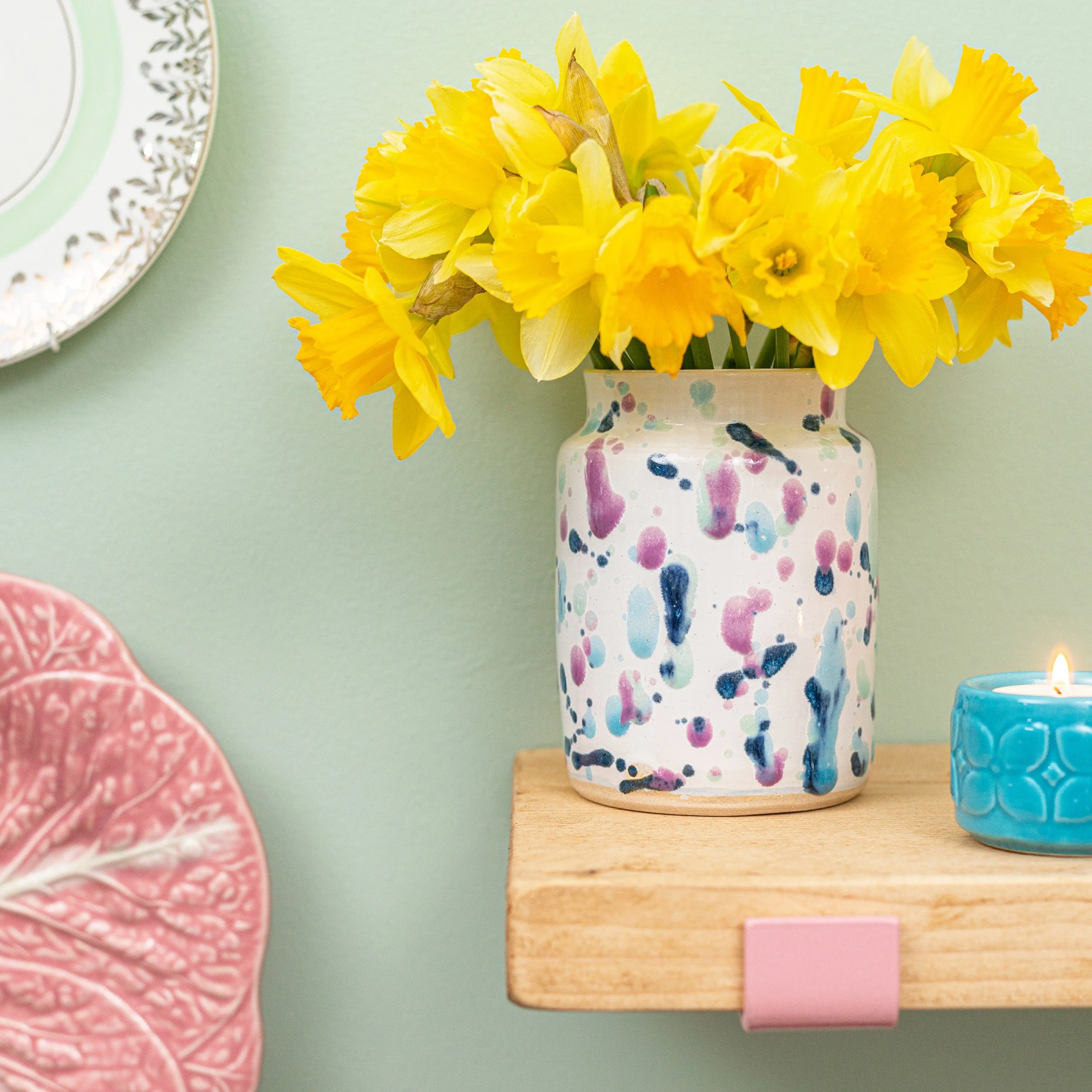 Yellow daffodils in a multicolored speckled ceramic vase on a wooden shelf, next to a lit blue candle and pink decorative plate against a light green wall.