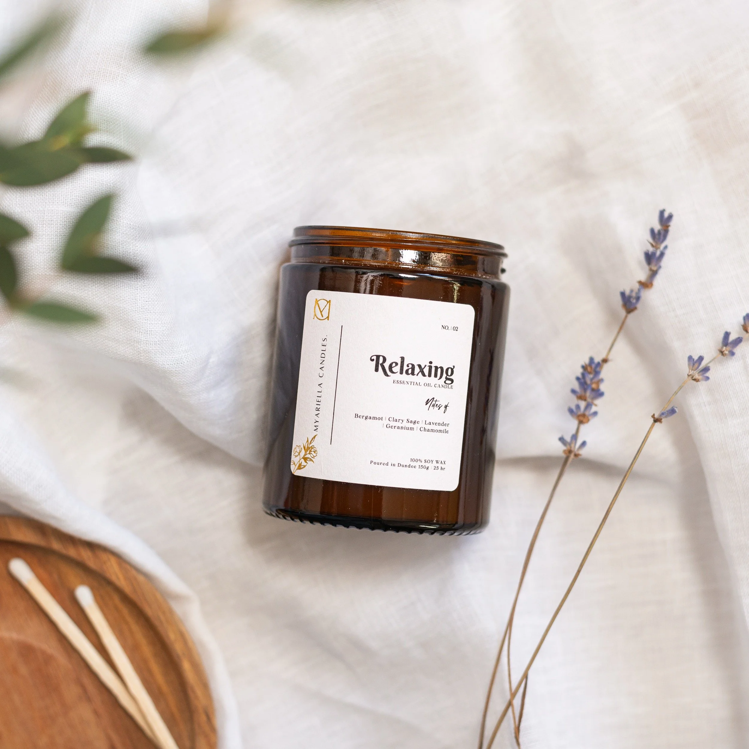 A brown glass candle jar labeled 'Relaxing' with essential oils, surrounded by dried lavender and greenery on white fabric.