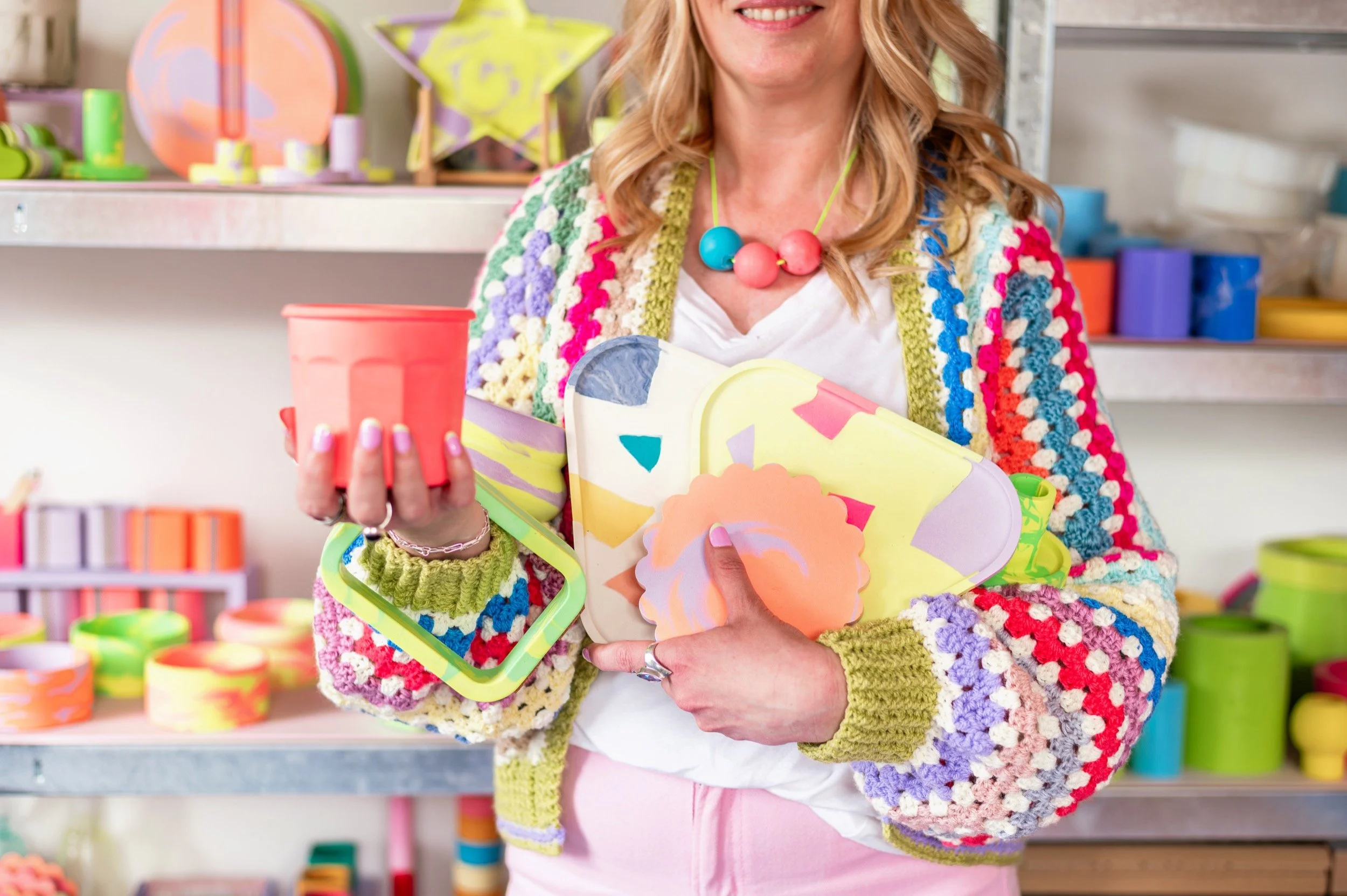 Woman holding colourful jesmonite pieces in her studio with shelves of colourful work behind her.