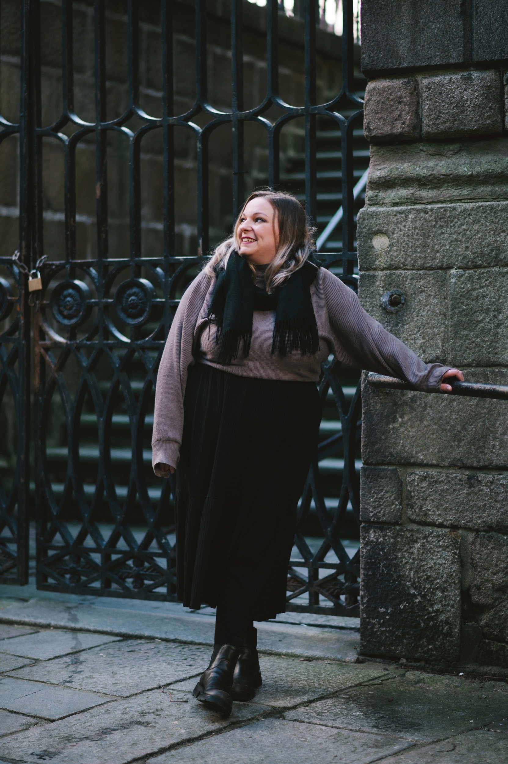 Ashleigh stands in front of a gothic gate, wearing dark toned clothes