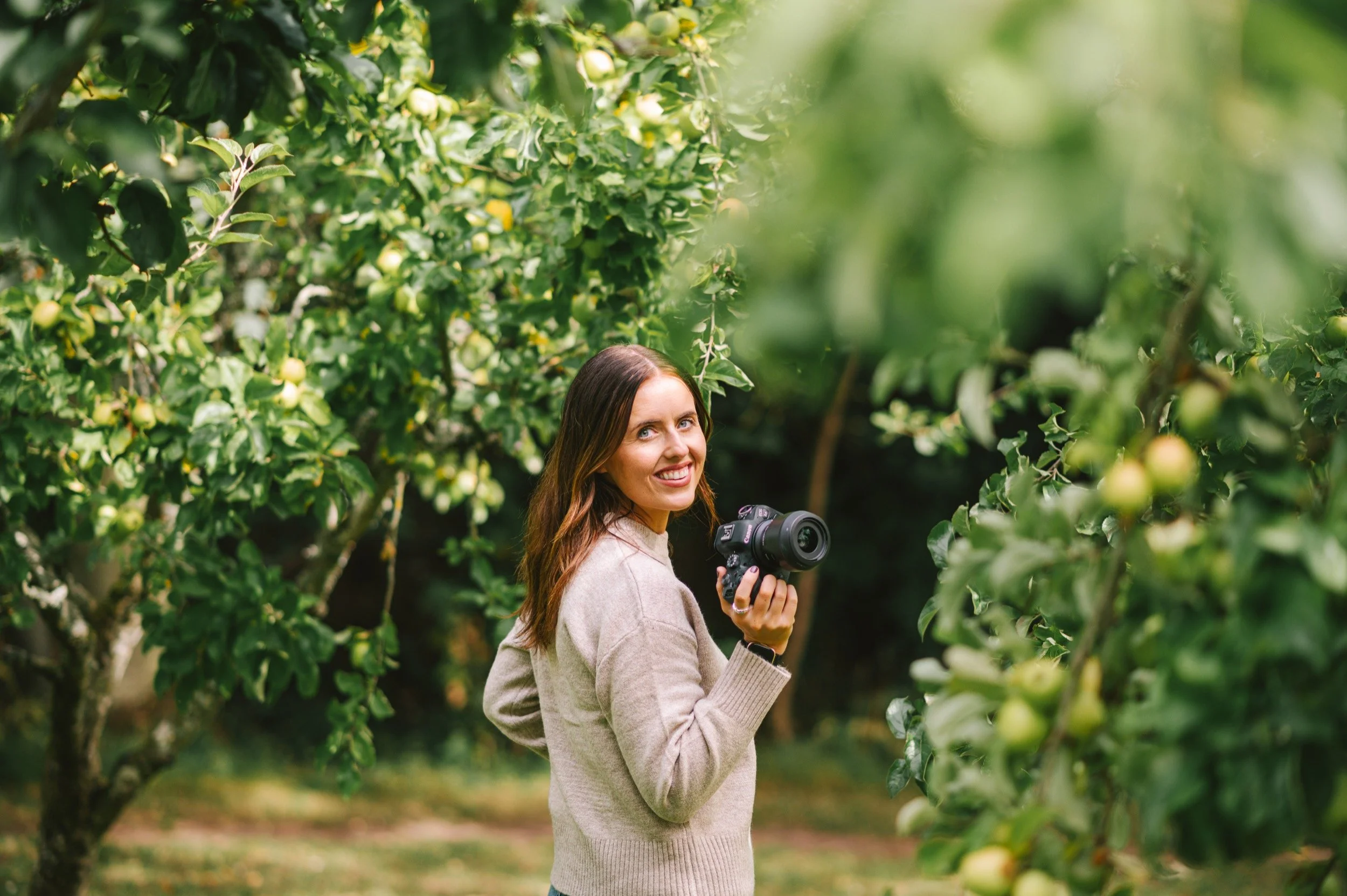 A woman in a beige sweater holding a camera stands among apple trees in an orchard, smiling at the camera.
