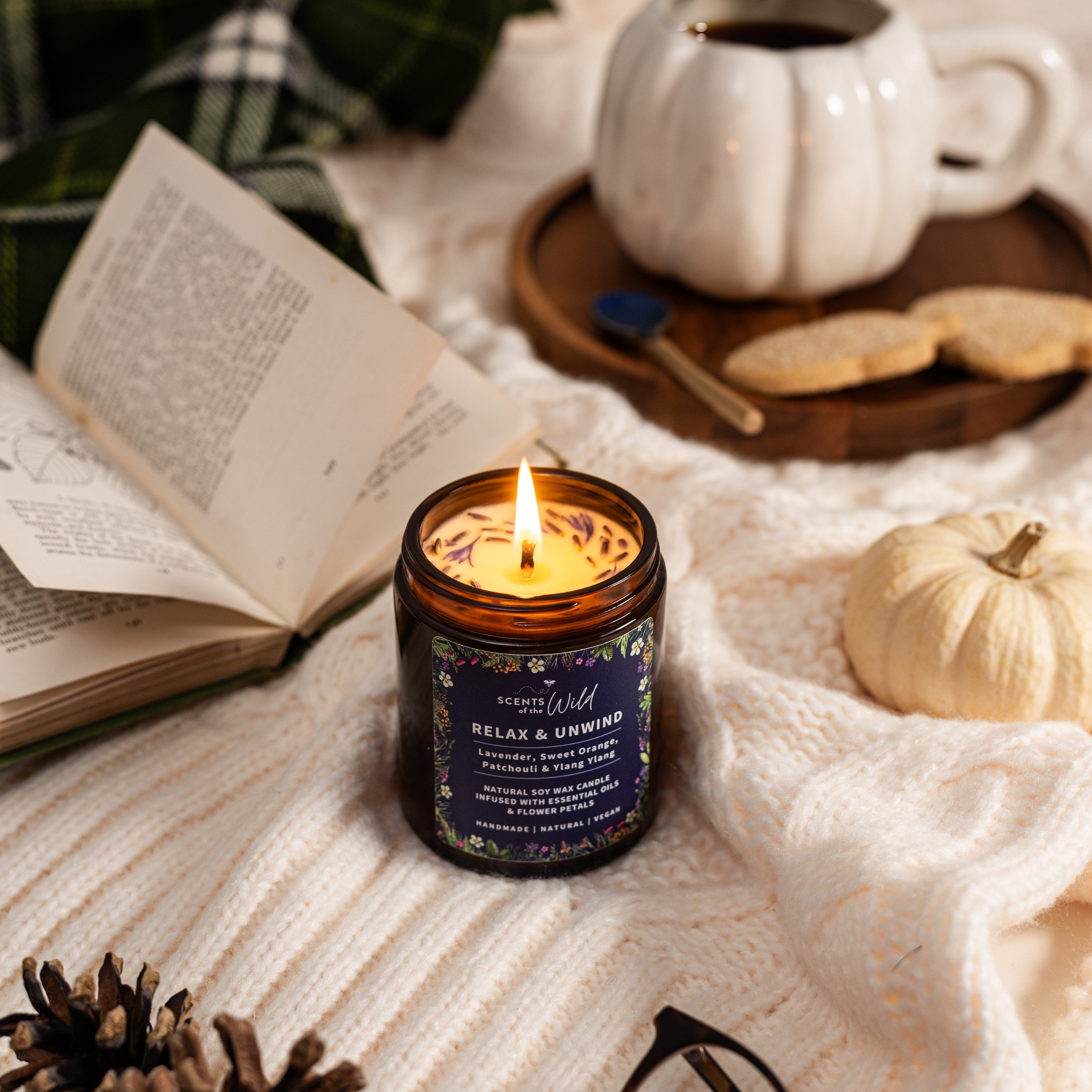 A cozy scene with a lit candle, an open book, a white pumpkin, a mug of coffee, and cookies on a wooden tray, all arranged on a soft cream-colored blanket.