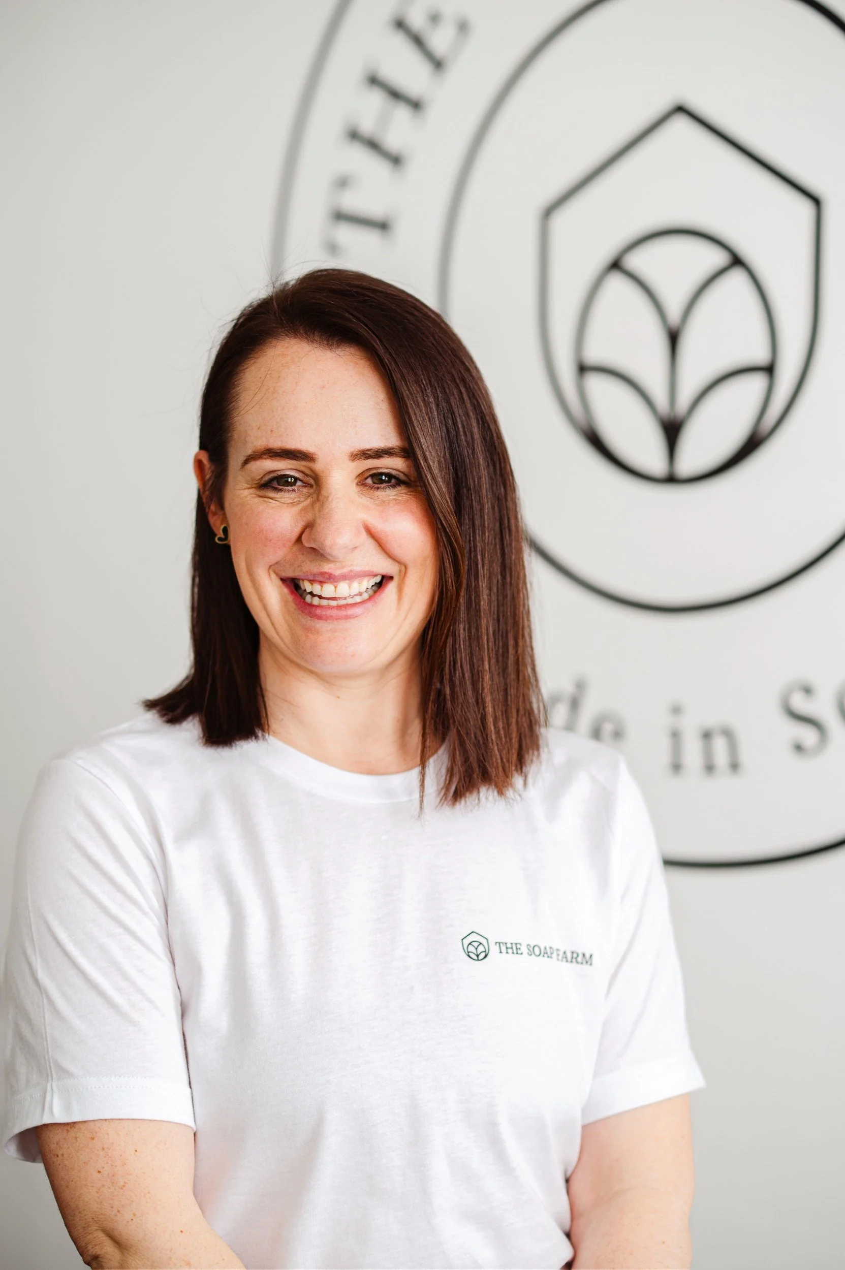 A smiling woman with shoulder-length brown hair wearing a white t-shirt with the logo 'The Soap Farm' standing in front of a wall with a logo and partial text.