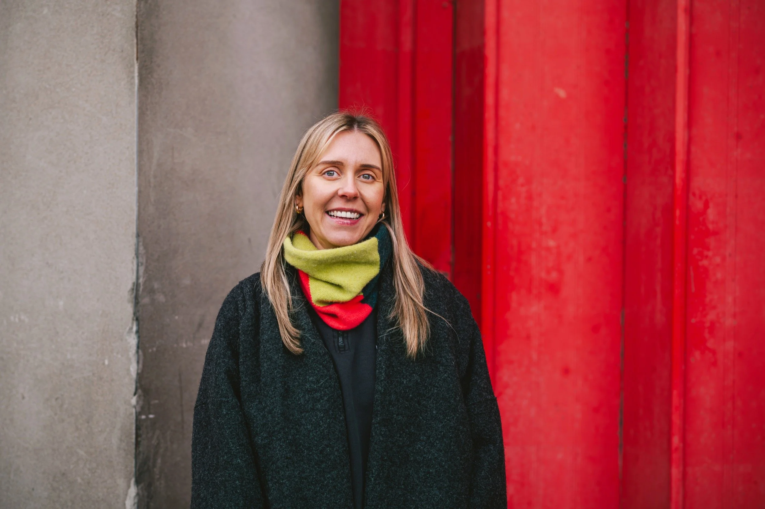 A woman with blonde hair smiling outdoors, wearing a black coat and a green, red, and navy snood, standing in front of a textured gray wall and a large red wooden door.